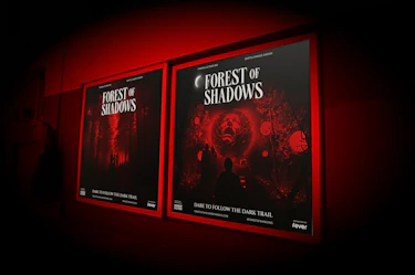 Backlit promotional posters displayed along a wall, featuring the title “FOREST OF SHADOWS,” the location “SEATTLE CHINESE GARDEN,” and the tagline “DARE TO FOLLOW THE DARK TRAIL.” The visuals depict a red-lit forest, visitor silhouettes, and a glowing sphere revealing a spectral face. The posters also include the mentions “Moment Factory” and “fever,” within the Forest of Shadows experience by Moment Factory.