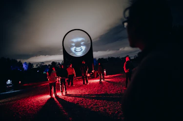 A group of people gathered in the evening along a path bathed in red light, facing a monumental circular projection displaying a glowing face, at the heart of an immersive installation within the Forest of Shadows experience by Moment Factory.