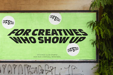 Bright green poster on a wall reading “For creatives who show up” in bold black distorted typography, with multiple “Offstage” logos in white circles. Location details below: Offstage Club Sportif, 5420 Rue Chapleau, Montréal. Surrounded by a worn wall, graffiti, and leafy plants.