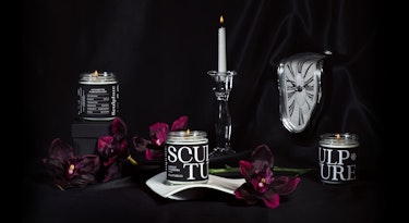 An image of three Sculpture branded candles displayed on a black background, where we can see a melting clock in the style of Salvador Dalí, a lit candle in a tall glass candle holder, and dark magenta flowers. 