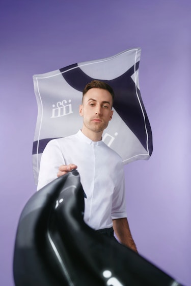 Karim Marier-El Khayat, cofondateur de Sculpture, debout devant un fond violet avec un morceau de tissu affichant le logo de Sculpture, portant une chemise blanche et un pantalon noir, agitant un morceau de tissu noir vers la caméra.