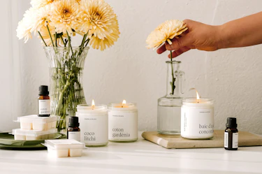 Marée soy candles in the scents Coco Litchi, Coton Gardénia, and Baie d'Açaï Concombre displayed with matching fragrance oils and wax melts beside fresh flowers, as a hand arranges a bouquet in a glass vase.