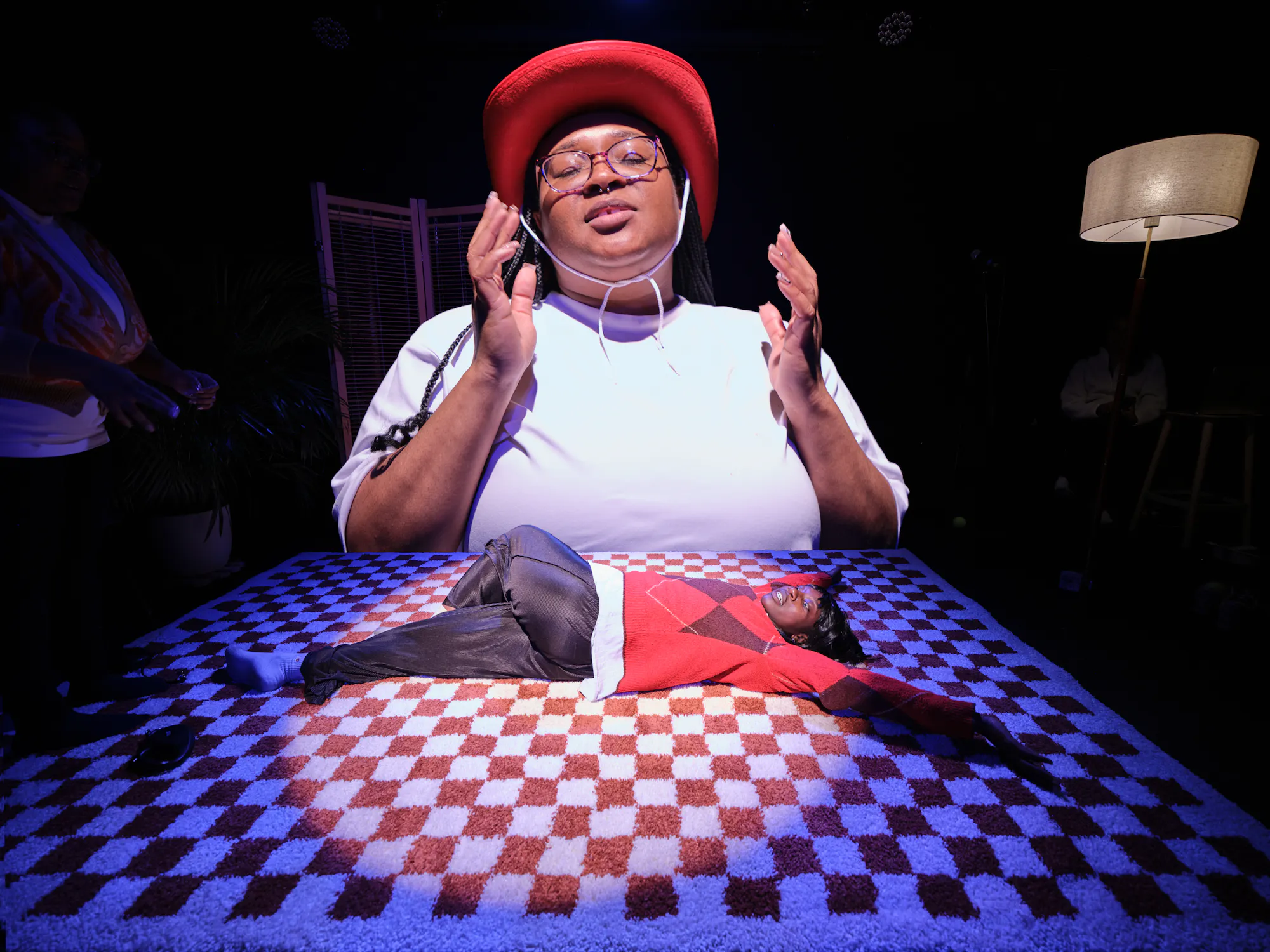 A black woman wearing glasses, a white t shirt and a red cowboy hat, has her eyes closed and hands either side of her face. In front of her is another black woman wearing a red jumper and dark grey trousers lying on her back on top of a checkerboard rug with her eyes closed. The image is a digital composite of two separate images, the woman in the cowboy hat is much bigger comparatively than the woman on the rug, making her look like she is looming over the woman on the floor.