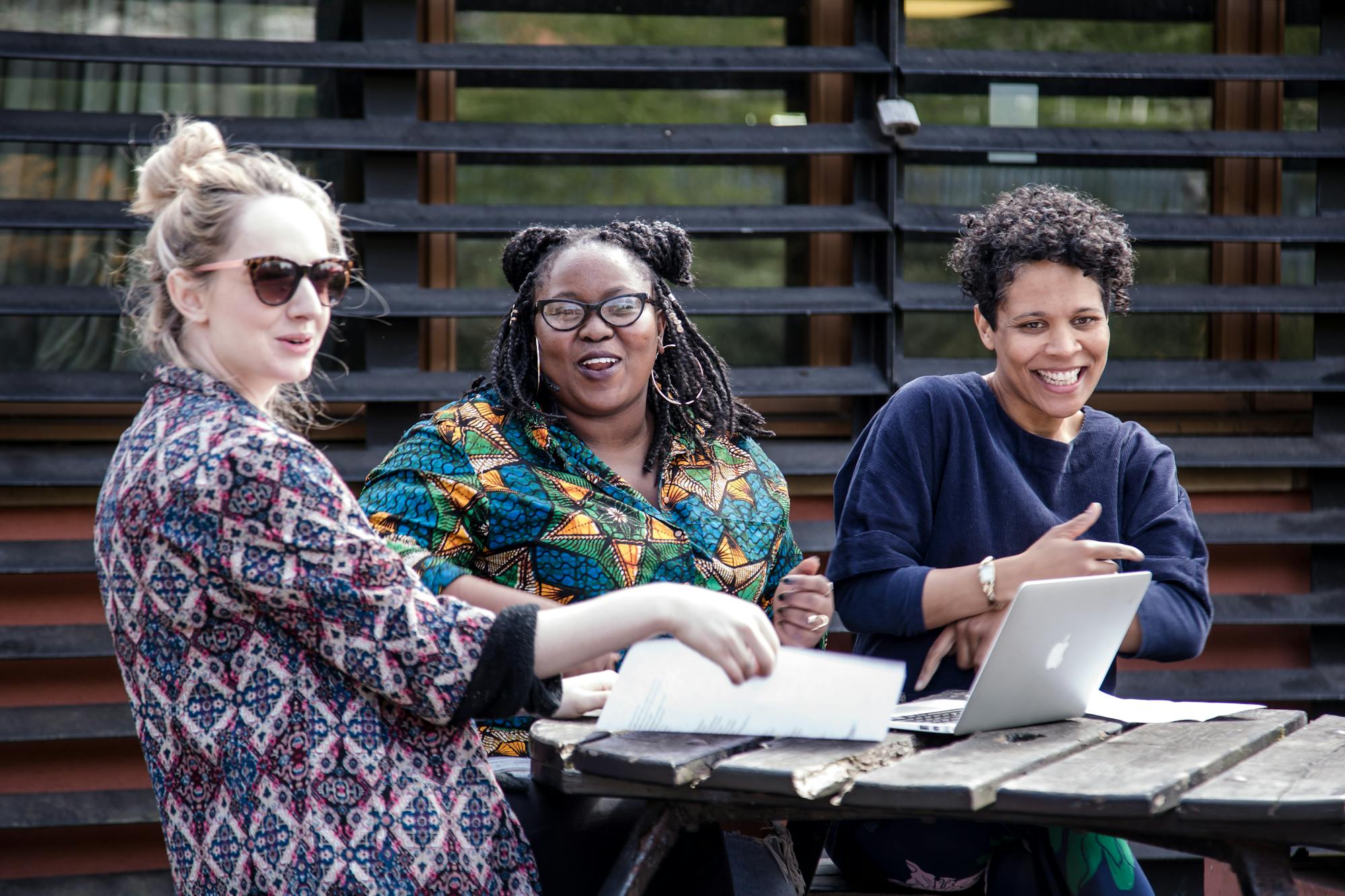 Three young women, one white and two black, sitting at an outdoor table, with notes and a laptop. They're smiling and laughing.
