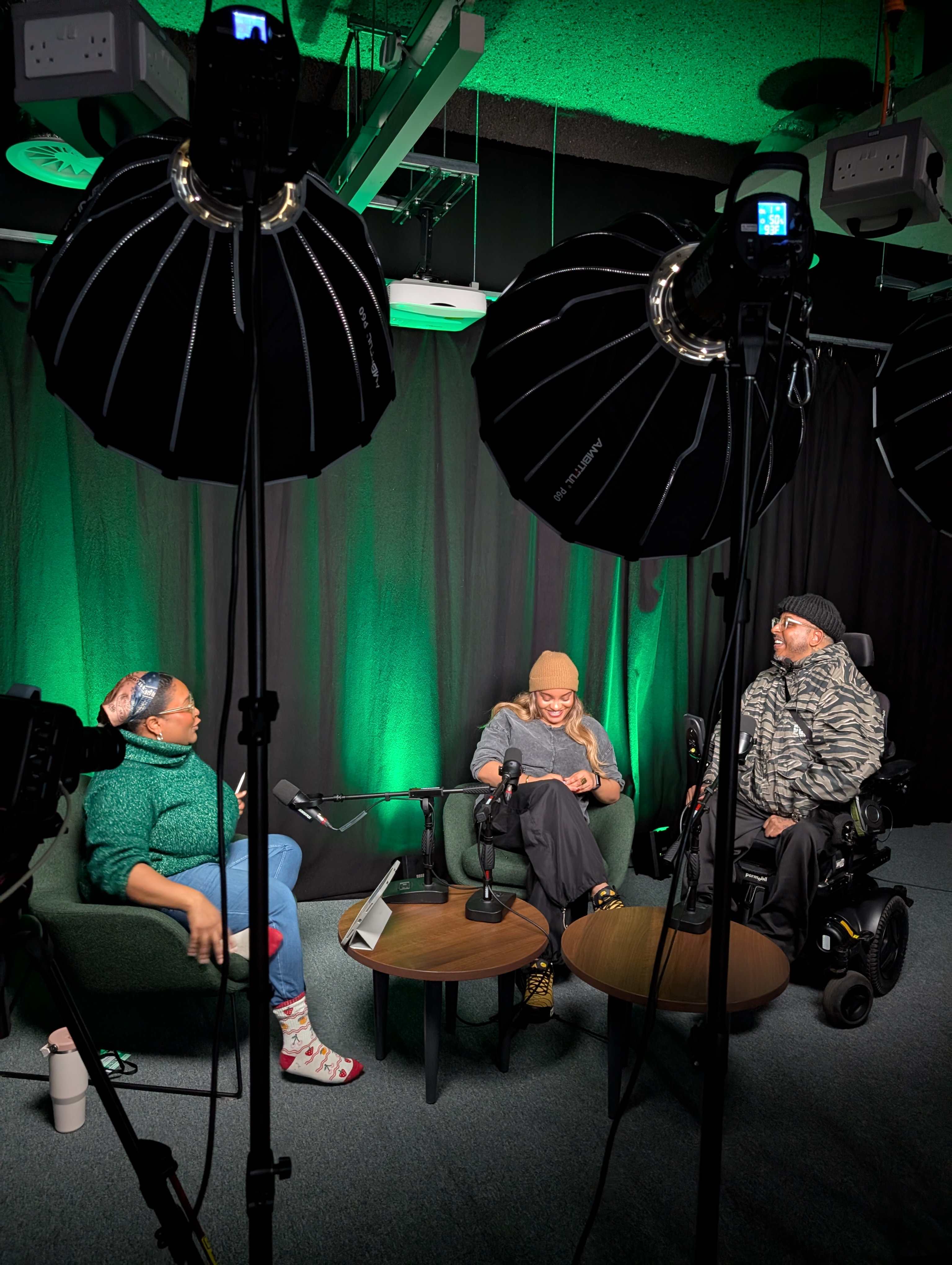 Three people sit in a podcast studio. They are in conversation with each other, in front of spotlights and microphones. The person on the right sits in a motorised wheelchair, with the other two people sat in armchairs.