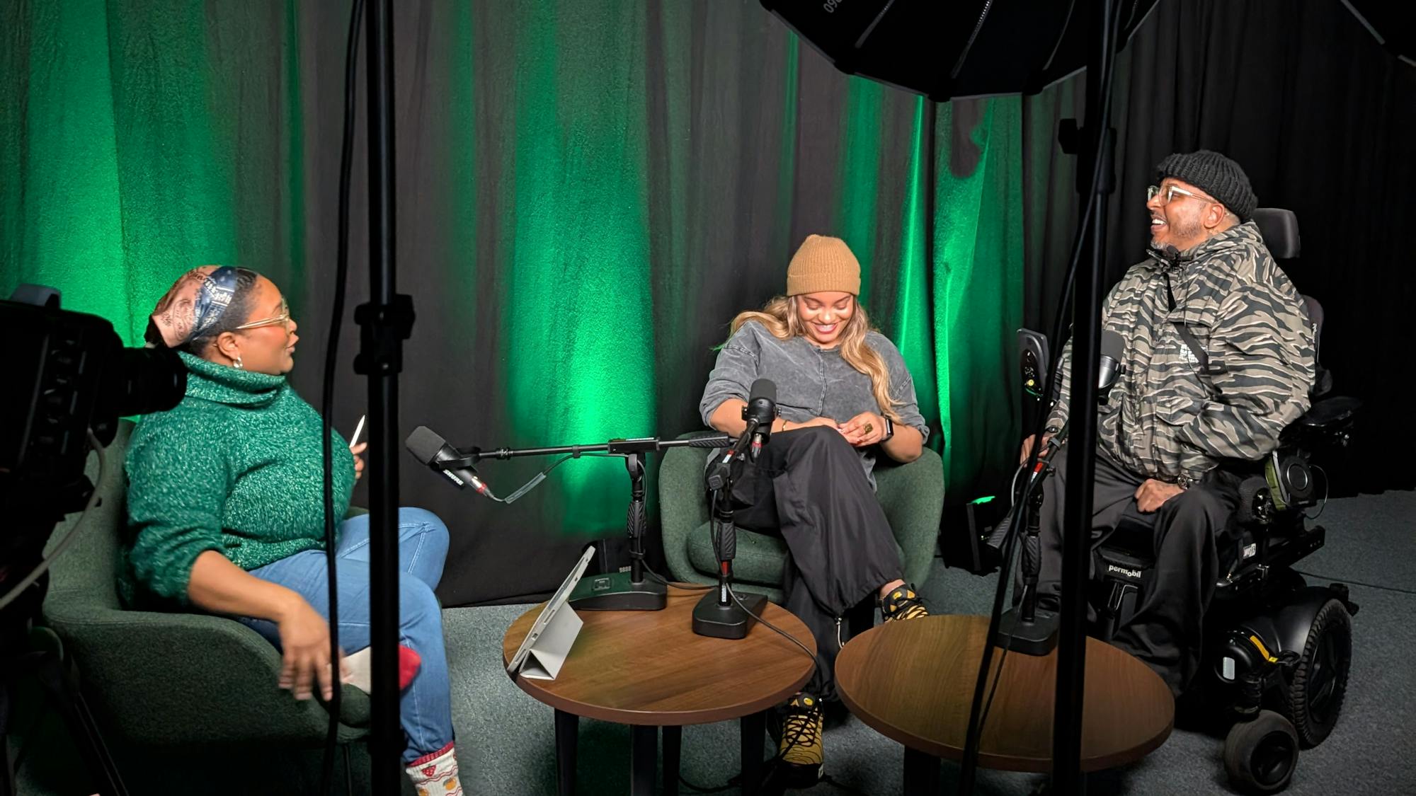 Three people sit in a podcast studio. They are in conversation with each other, in front of spotlights and microphones. The person on the right sits in a motorised wheelchair, with the other two people sat in armchairs.