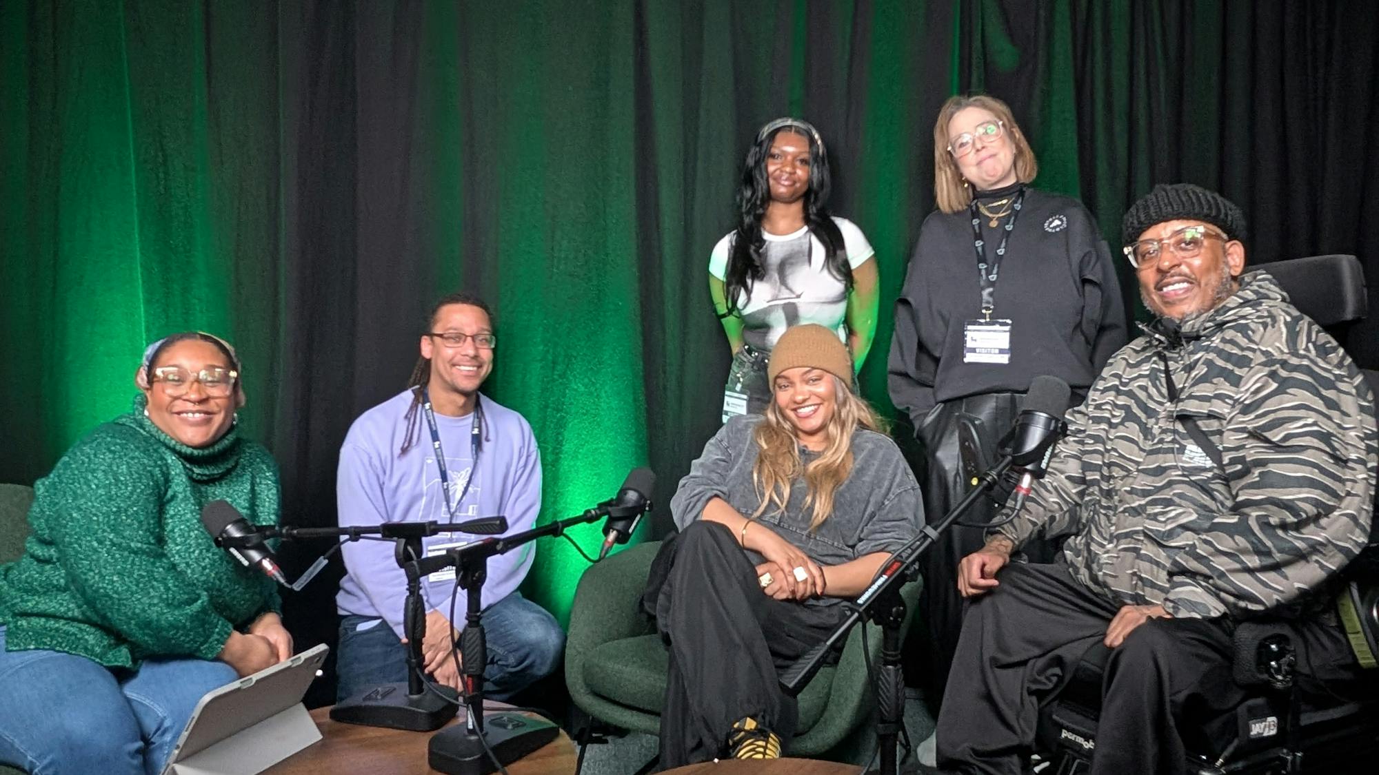 A group of six people smile to camera. They are in a podcast studio, in front of black drapes lit with green lighting. In front of them are two wooden coffee tables with microphones in stands resting on them. The four people at the front of the group are seated, with the person on the far right sat in a motorised wheelchair. The two people at the back are standing behind the seated people.