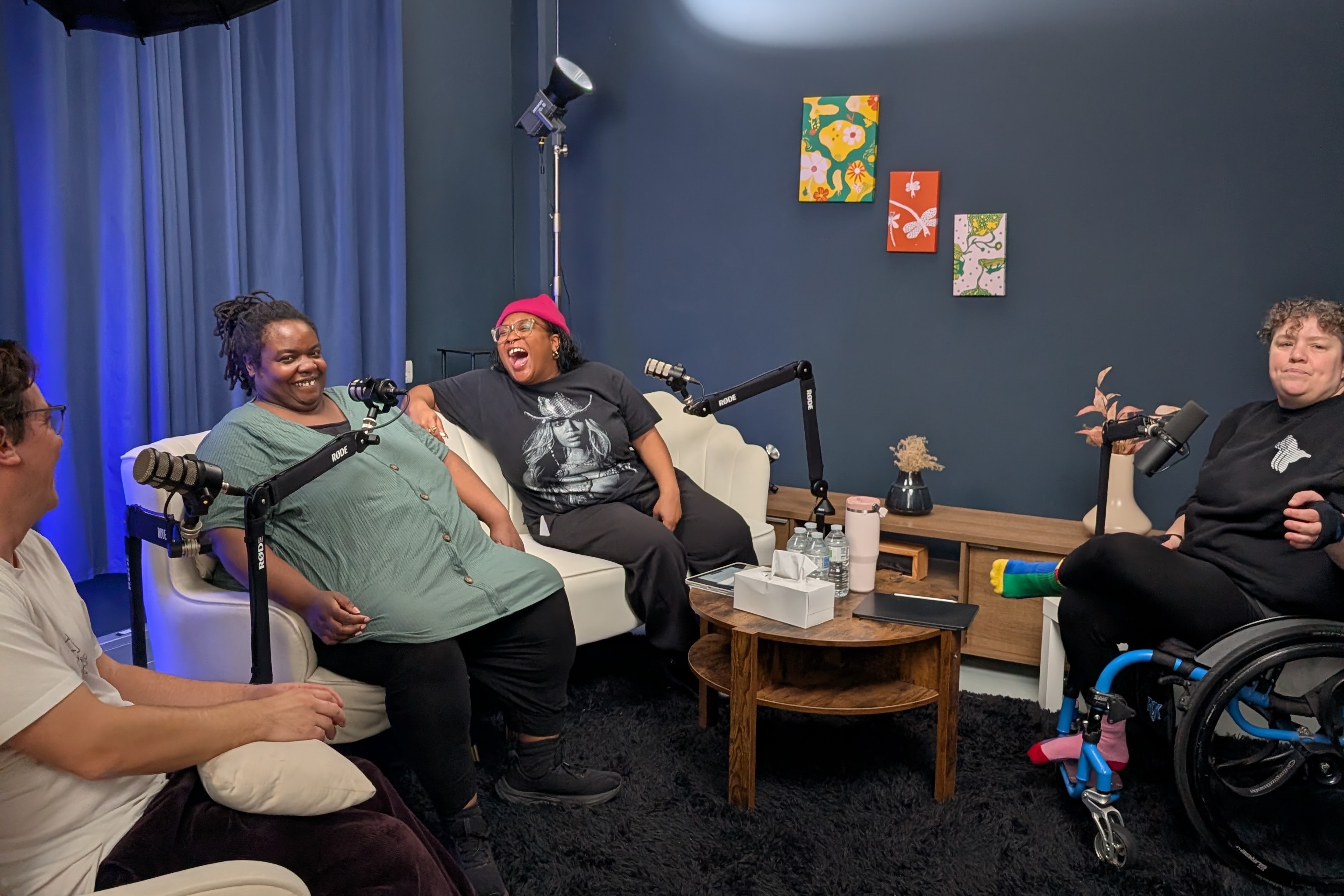 Four people sit in a room in conversation with each other. The person on the left sits on a chair, two people in the middle sit on a two-seater sofa, and a fourth person sits to the right in a wheelchair. There is a coffee table and two microphones in stands on floor between the seated people. The two people on the sofa are laughing.