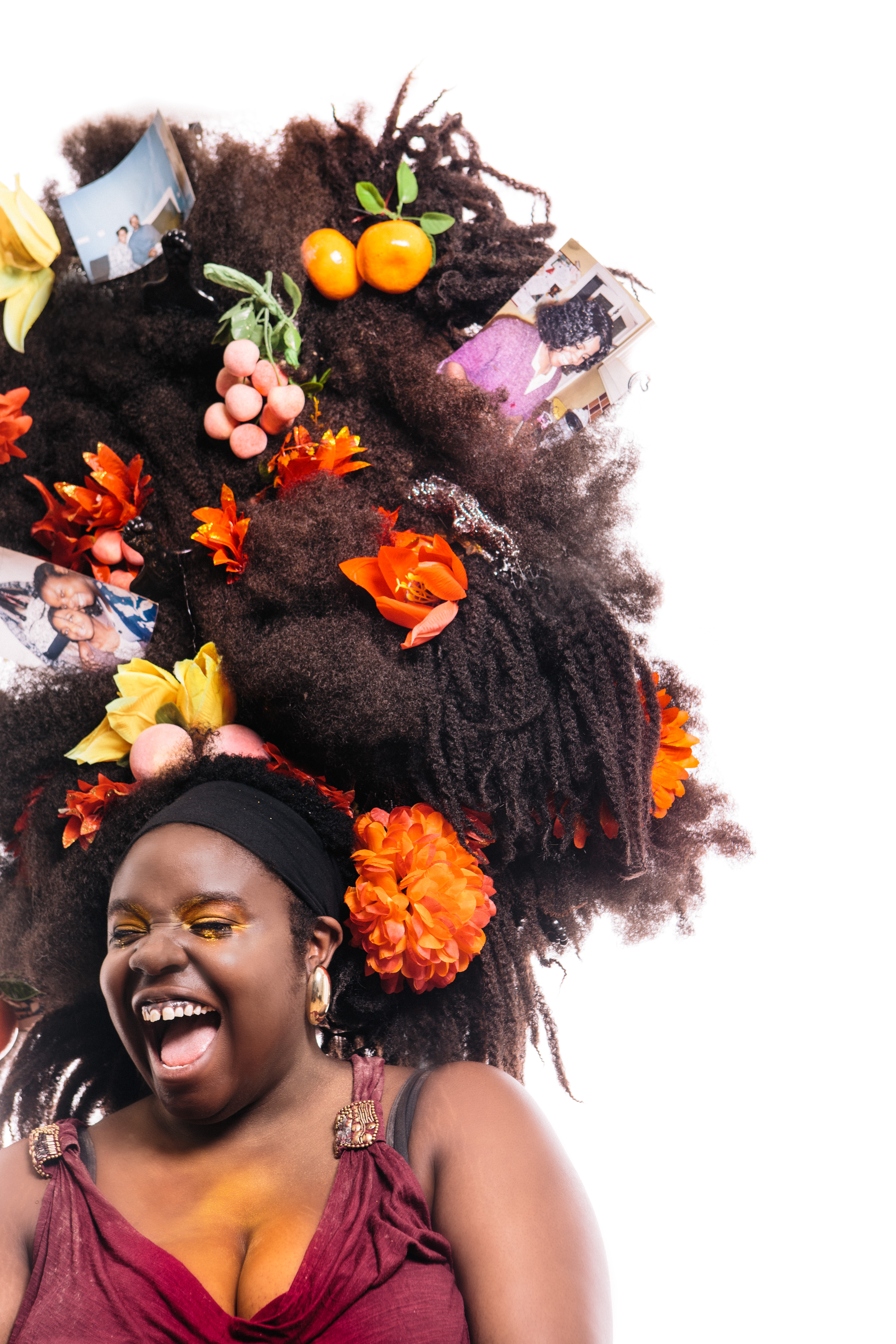 A black woman wearing a red dress and gold hoop earrings laughs. Her giant tumble-weave is filled with flowers, fruits and photographs.