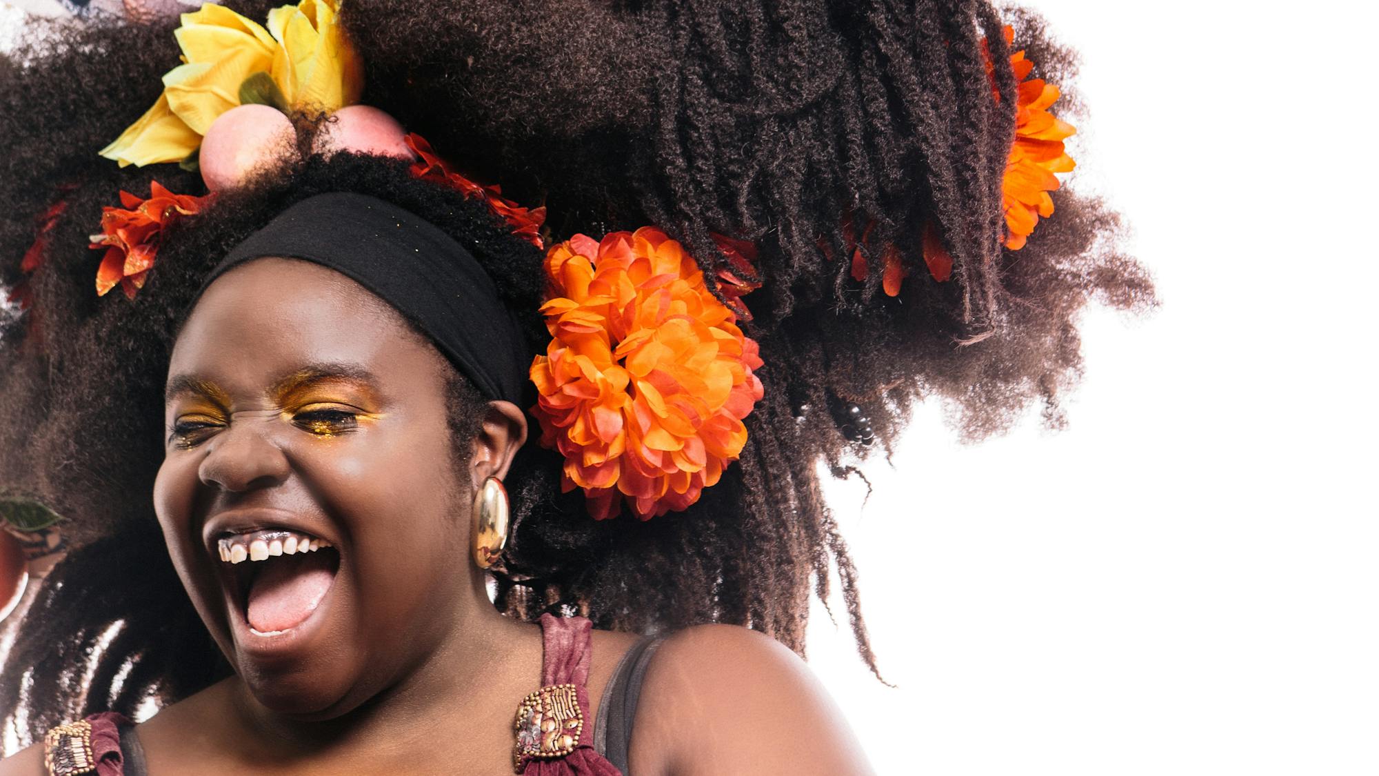 A black woman wearing a red dress and gold hoop earrings laughs. Her giant tumble-weave is filled with flowers, fruits and photographs.