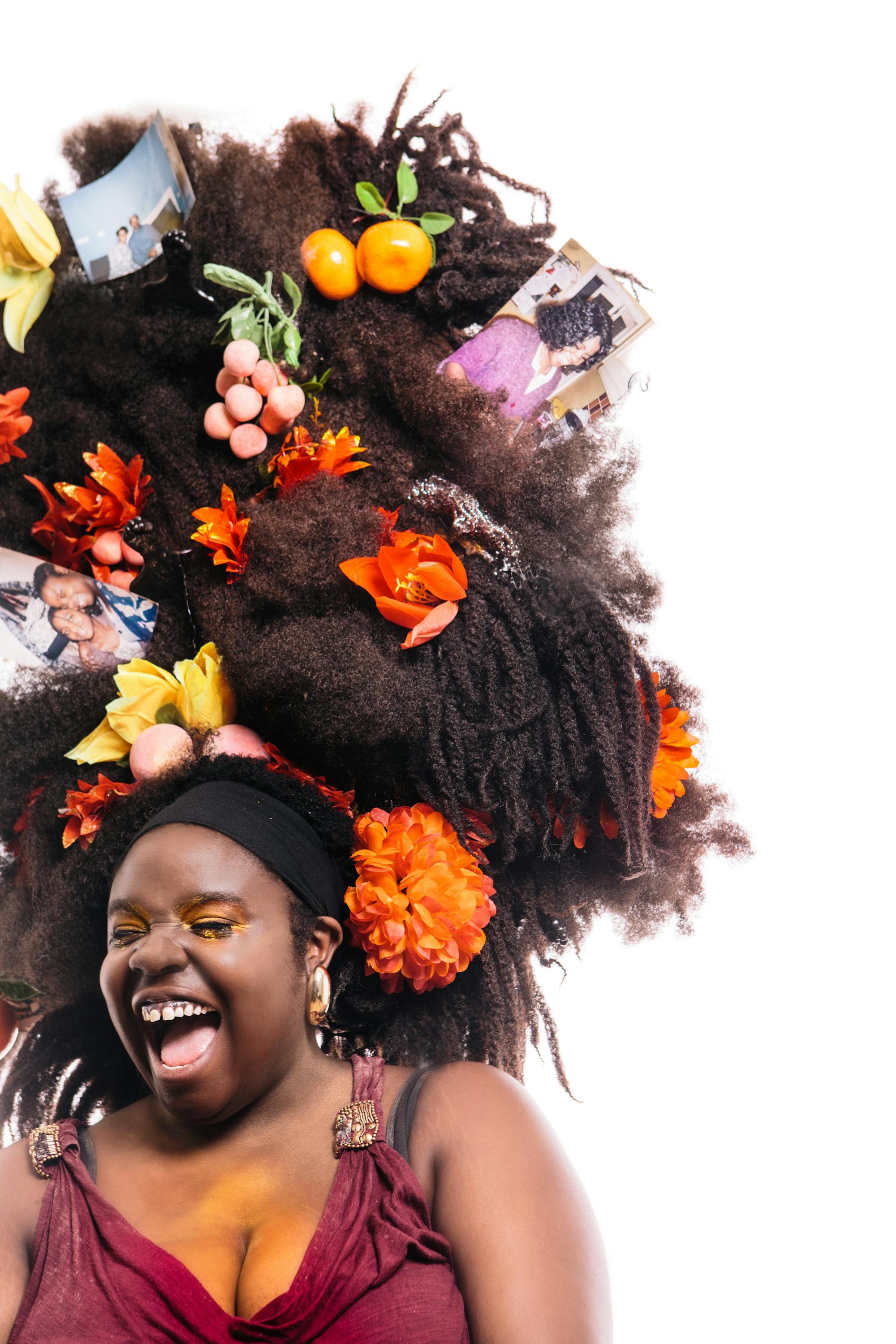 A black woman wearing a red dress and gold hoop earrings laughs. Her giant tumble-weave is filled with flowers, fruits and photographs.