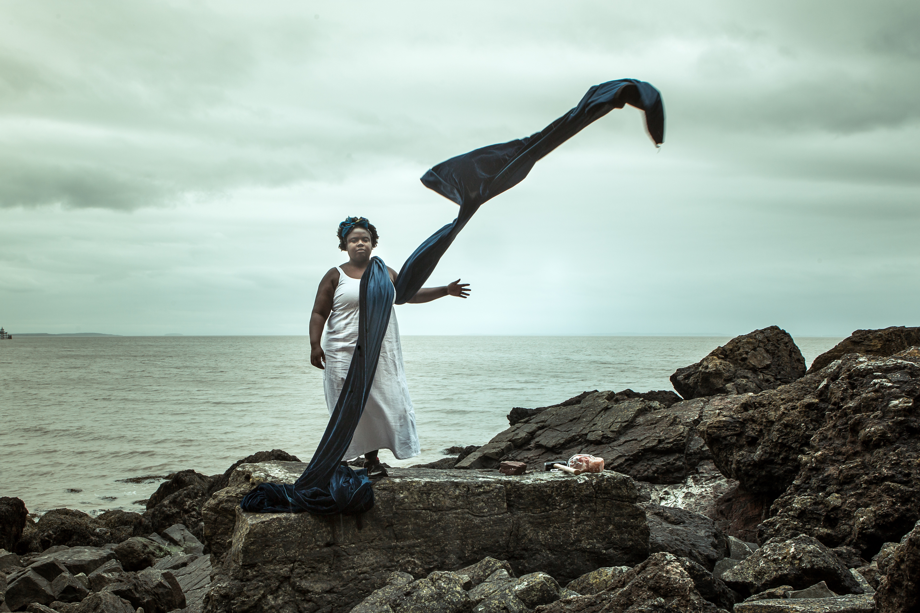 A black woman stands on rocks in front of the sea. The sky is grey and cloudy. She wears a very long blue scarf, which is blowing in the wind.