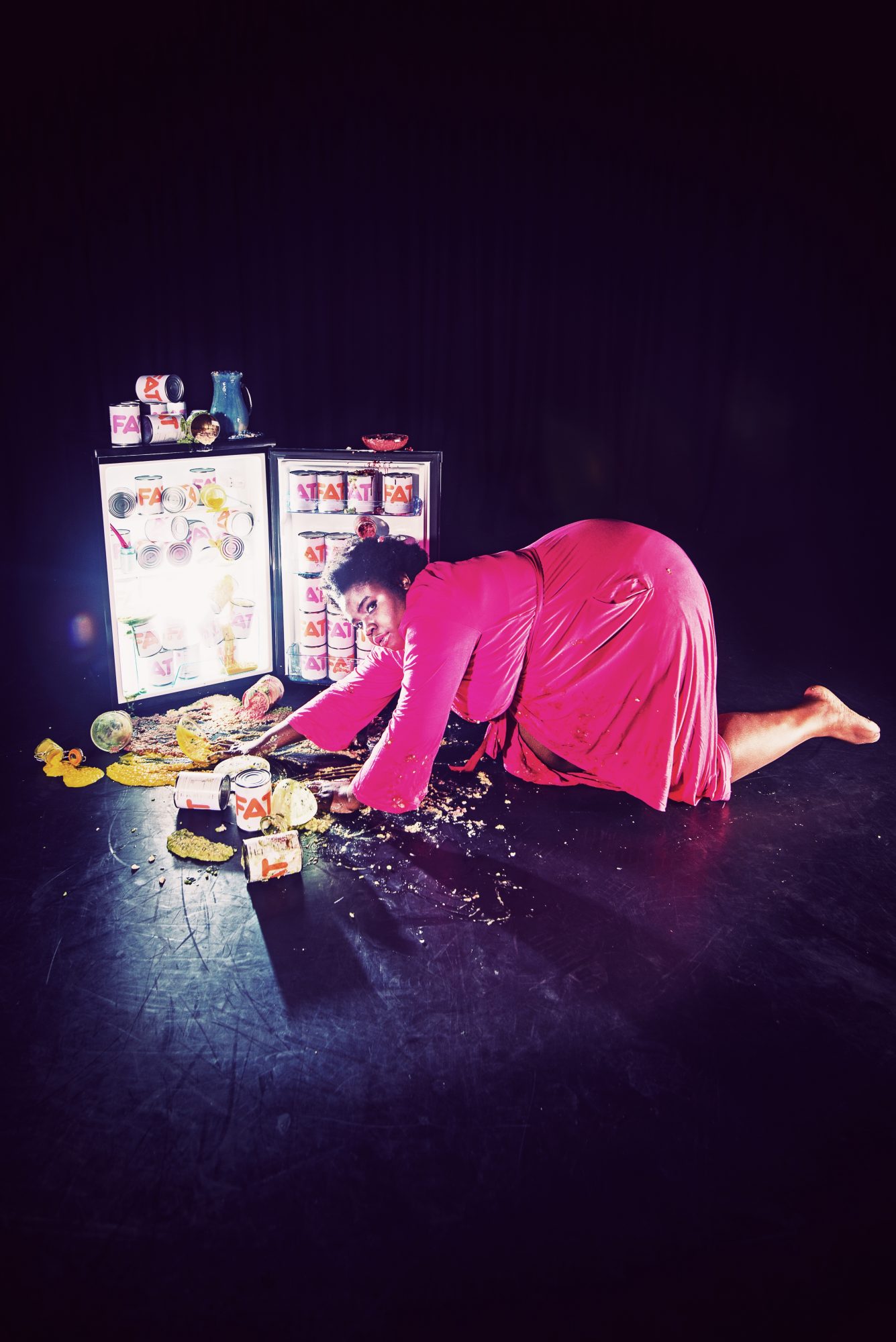 A black woman in a pink dress crawls across the floor toward an open fridge, illuminated from within and with food spilling out.