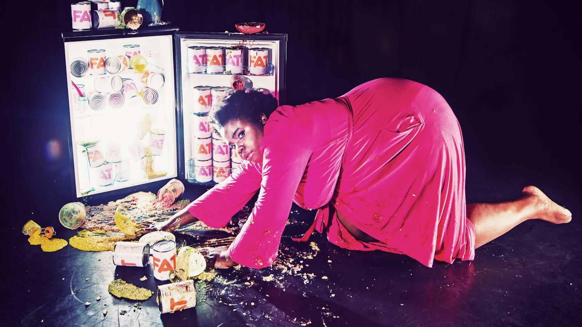 A black woman in a pink dress crawls across the floor toward an open fridge, illuminated from within and with food spilling out.