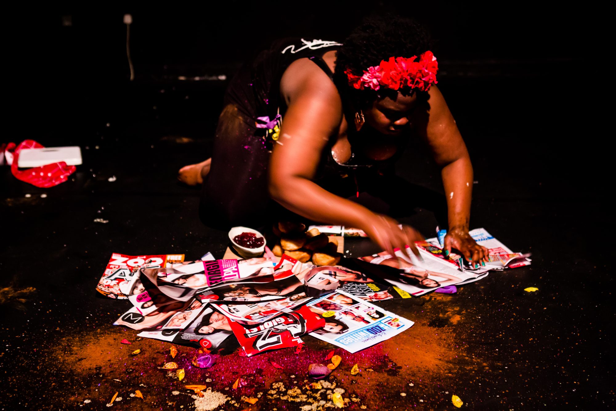 A black woman is on all fours, with several magazines spread out on the floor in front of her. She is poring through them.