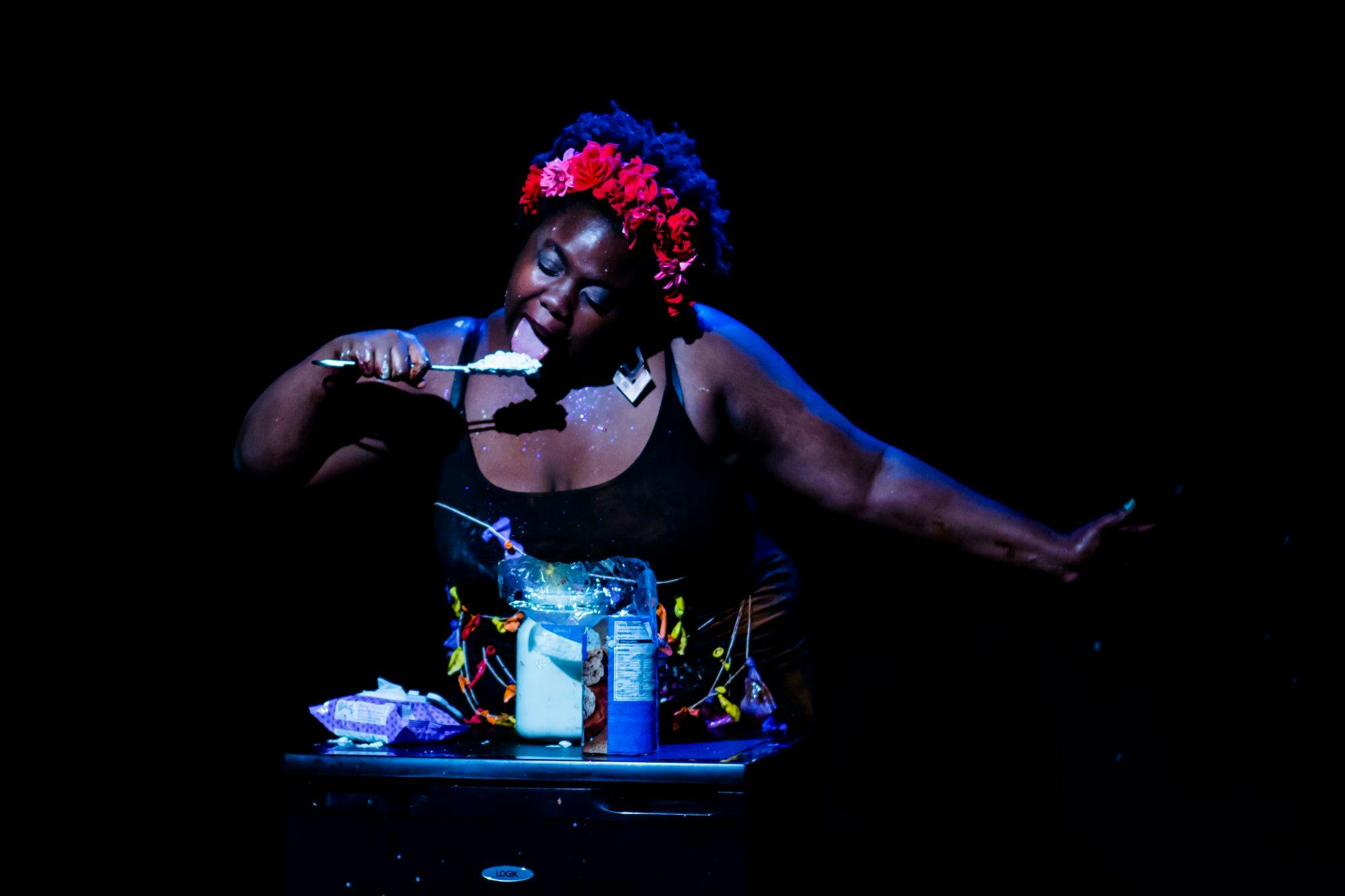 A black woman wearing a black dress and a red flower garland in her hair stands behind a table. On the table is a jar filled with rice pudding, and the woman is licking a spoon full of it.