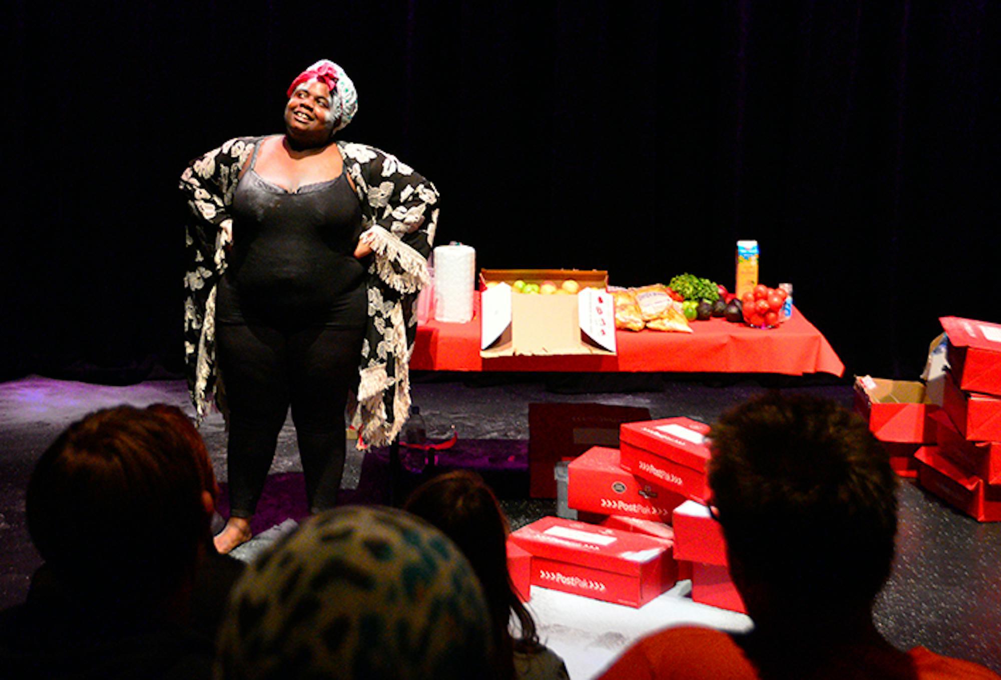 Selina, a Black woman, stands on stage, by a red table with boxes of food, and red postal boxes on the floor.