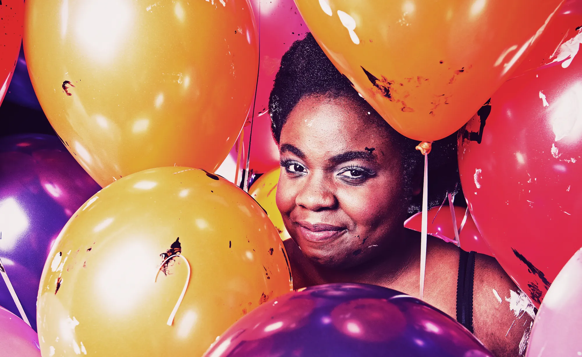 Selina, a black woman, smiles whilst surrounded by yellow, red and purple balloons. They obscure her body, so that you can only see her face and her left shoulder.
