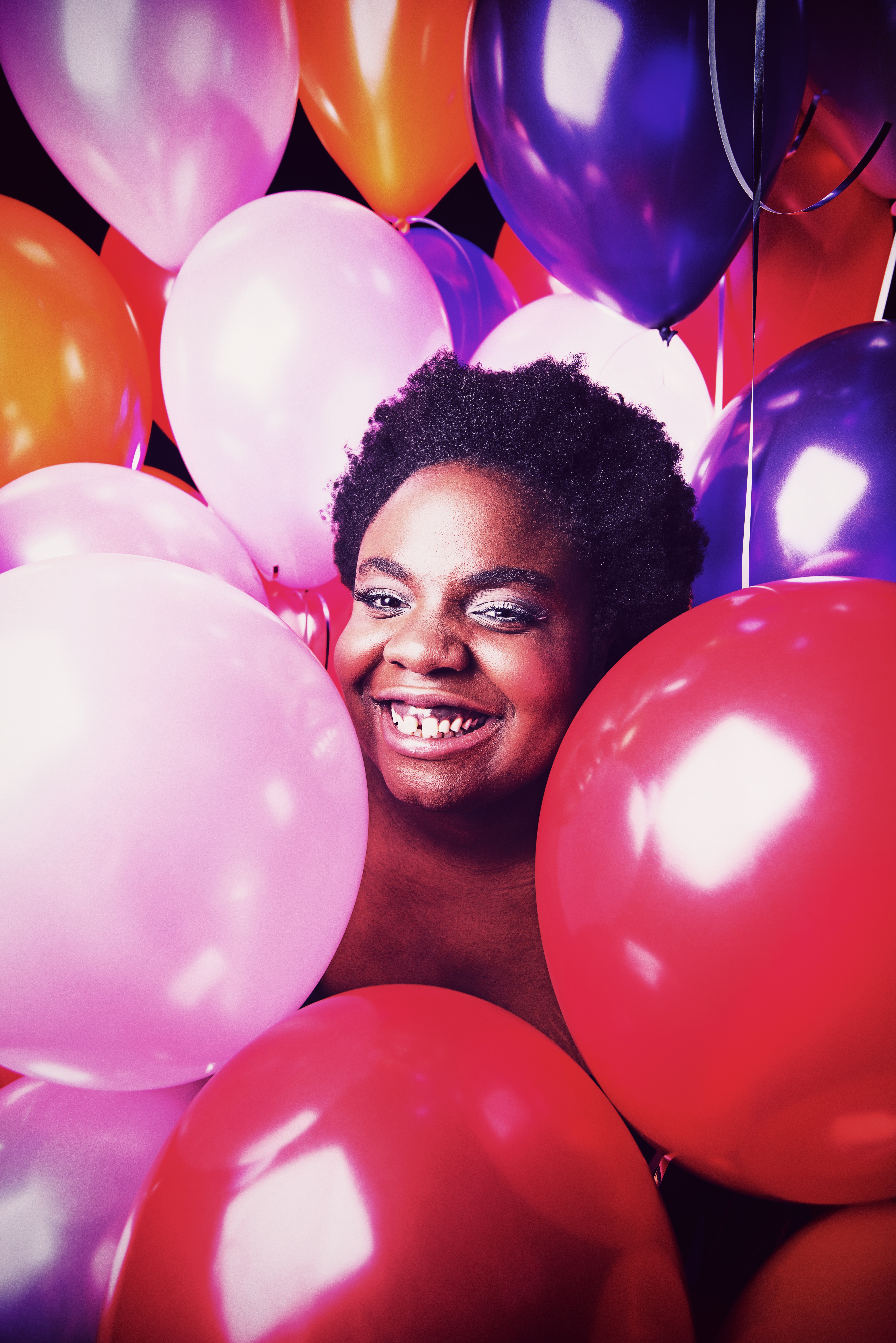 Selina, a black woman, smiles whilst surrounded by pink, red, purple snd orange balloons. They obscure her body, so that you can only see her face and her chest.