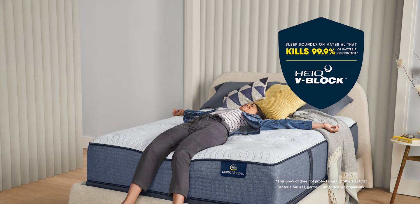 A woman lays on a Serta Perfect Sleeper mattress in a bedroom. A blue shield is overlaid on the image with text that reads: Sleep soundly on material that kills 99.9% of bacteria on contact. HeiQ V-Block. This product does not protect users or others against bacteria, viruses, germs or other disease organisms.