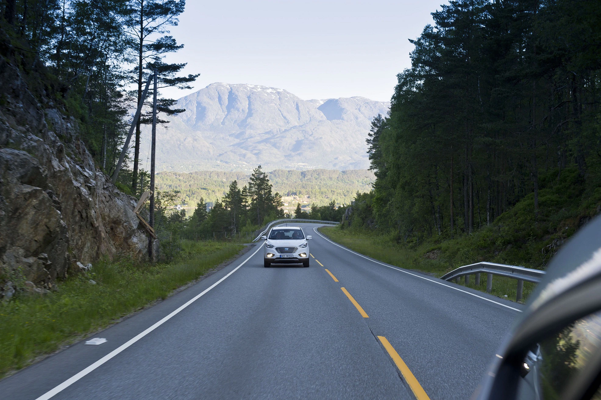 Auto fährt auf kurviger Landstraße durch bewaldete Gegend mit Bergen im Hintergrund und blauem Himmel.