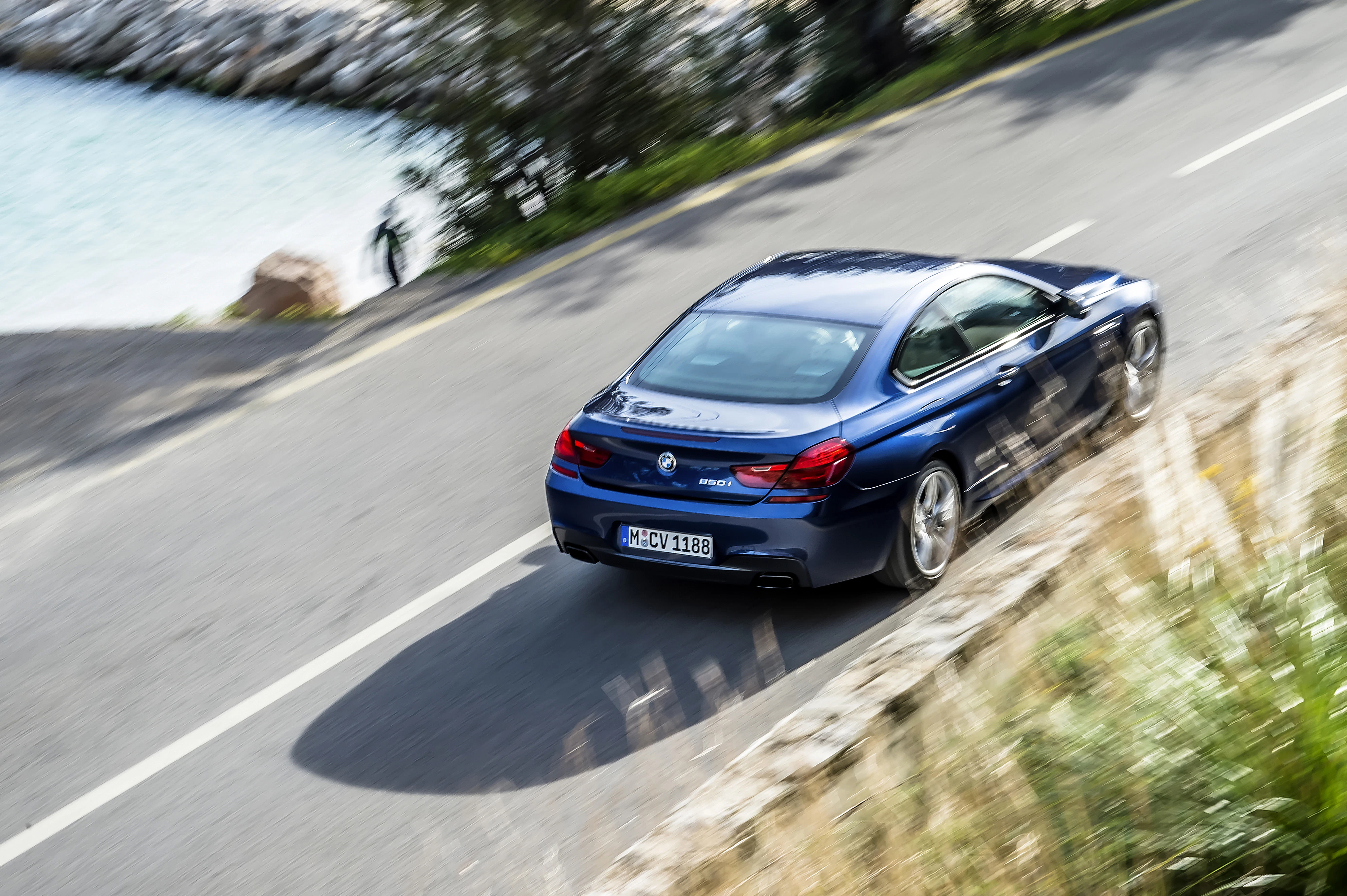 Blauer BMW fährt auf einer kurvigen Küstenstraße entlang des Meeres, umgeben von grüner Vegetation und sonnigem Wetter.