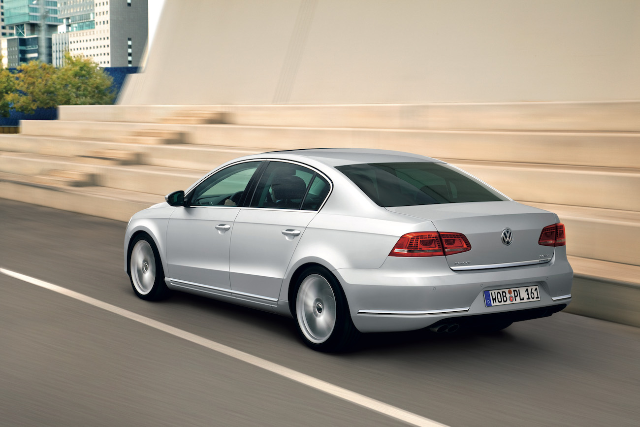 Silberner Volkswagen Passat fährt schnell auf einer Straße in urbaner Umgebung, moderne Gebäude im Hintergrund.