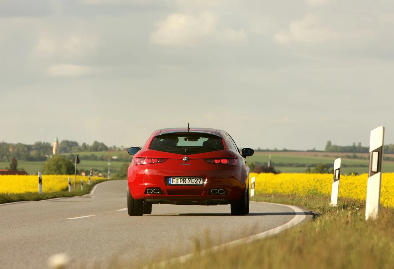 Ein rotes Auto fährt auf einer Landstraße durch eine ländliche Landschaft mit gelb blühenden Feldern und einem bewölkten Himmel im Hintergrund.