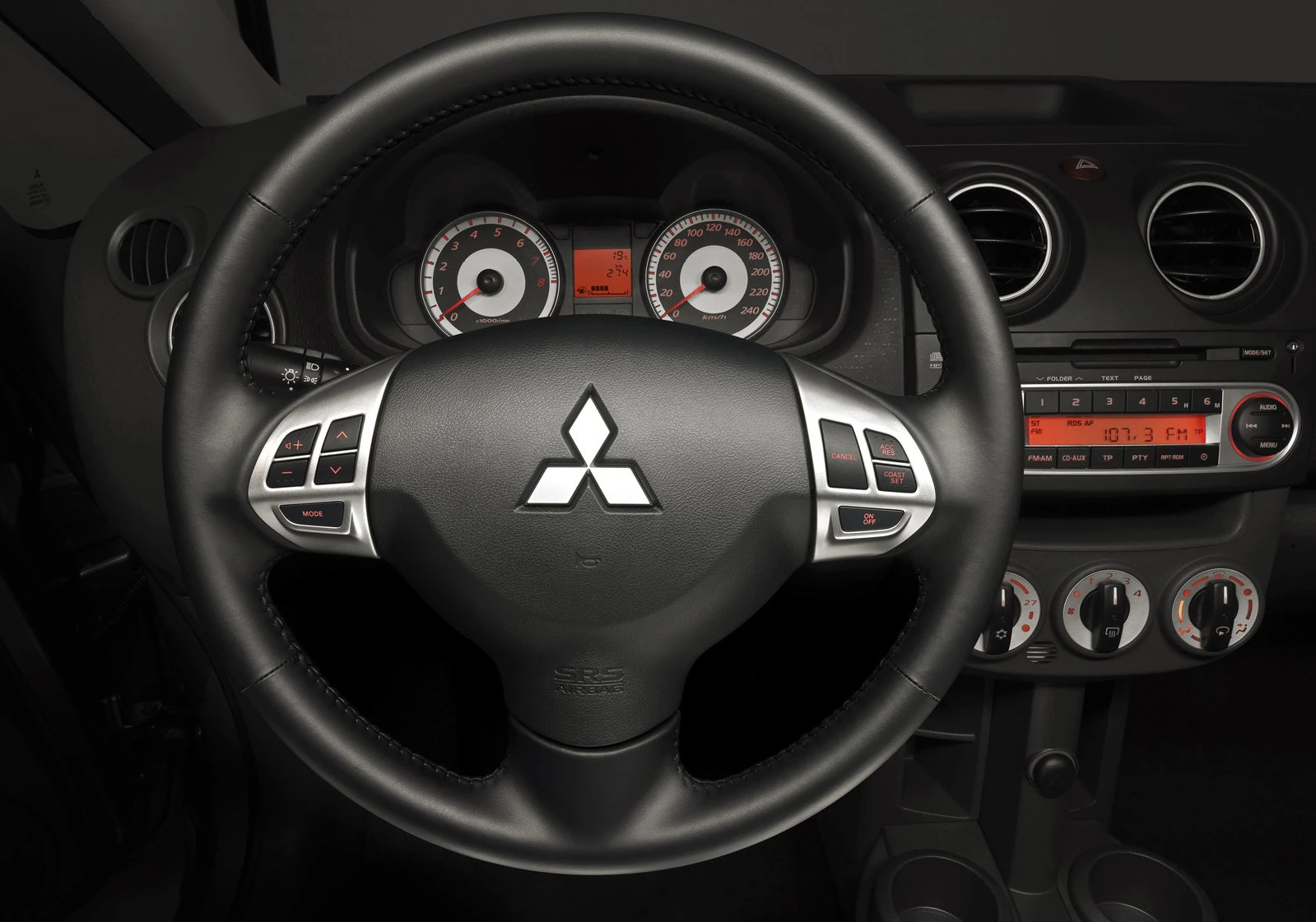 Lenkrad eines Mitsubishi-Autos mit Logo, rundem Armaturenbrett, Tachoanzeigen und Bedienelementen für Klima und Radio.