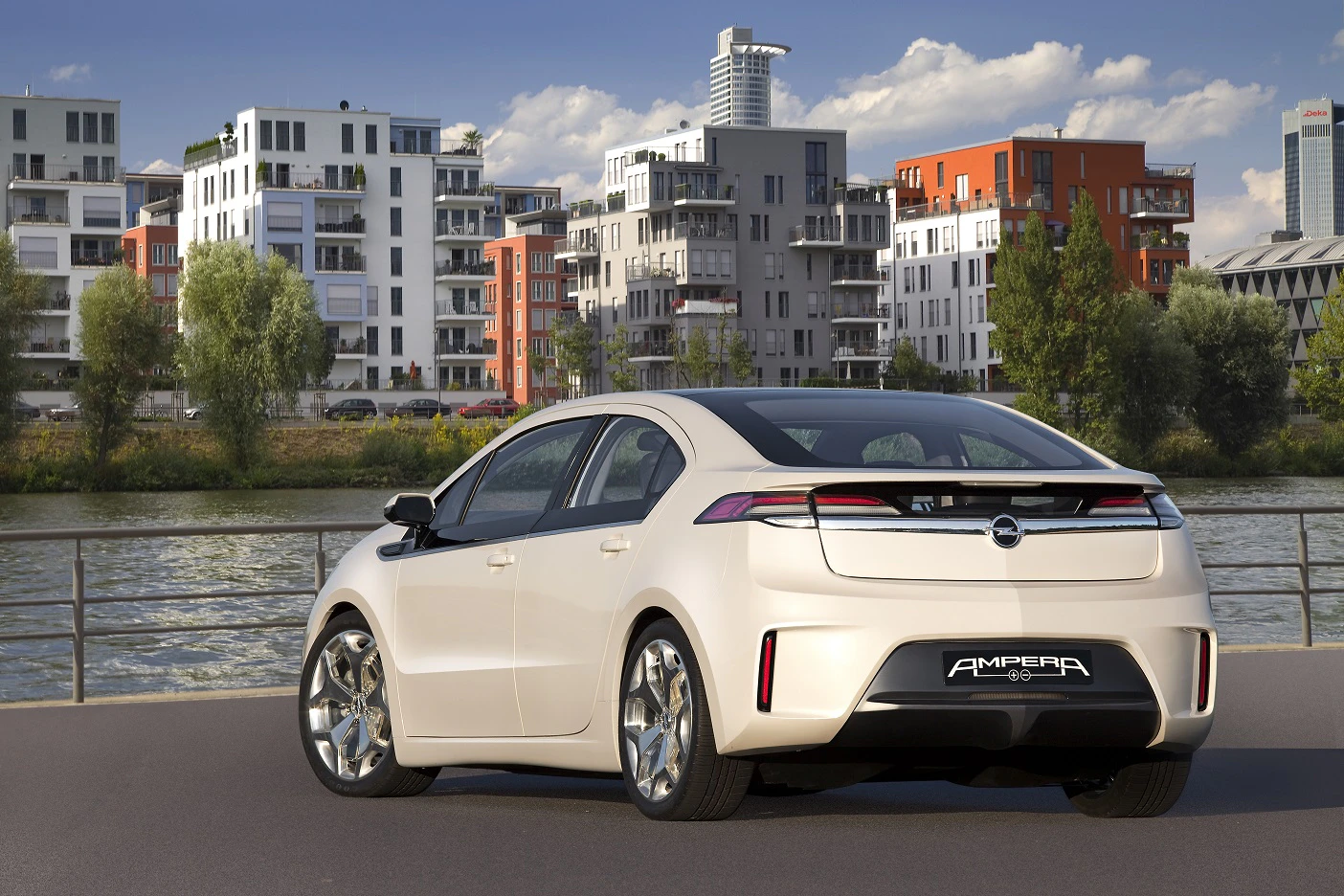 Weißer Opel Ampera geparkt an einem Fluss, moderne Gebäude im Hintergrund, sonniger Himmel.