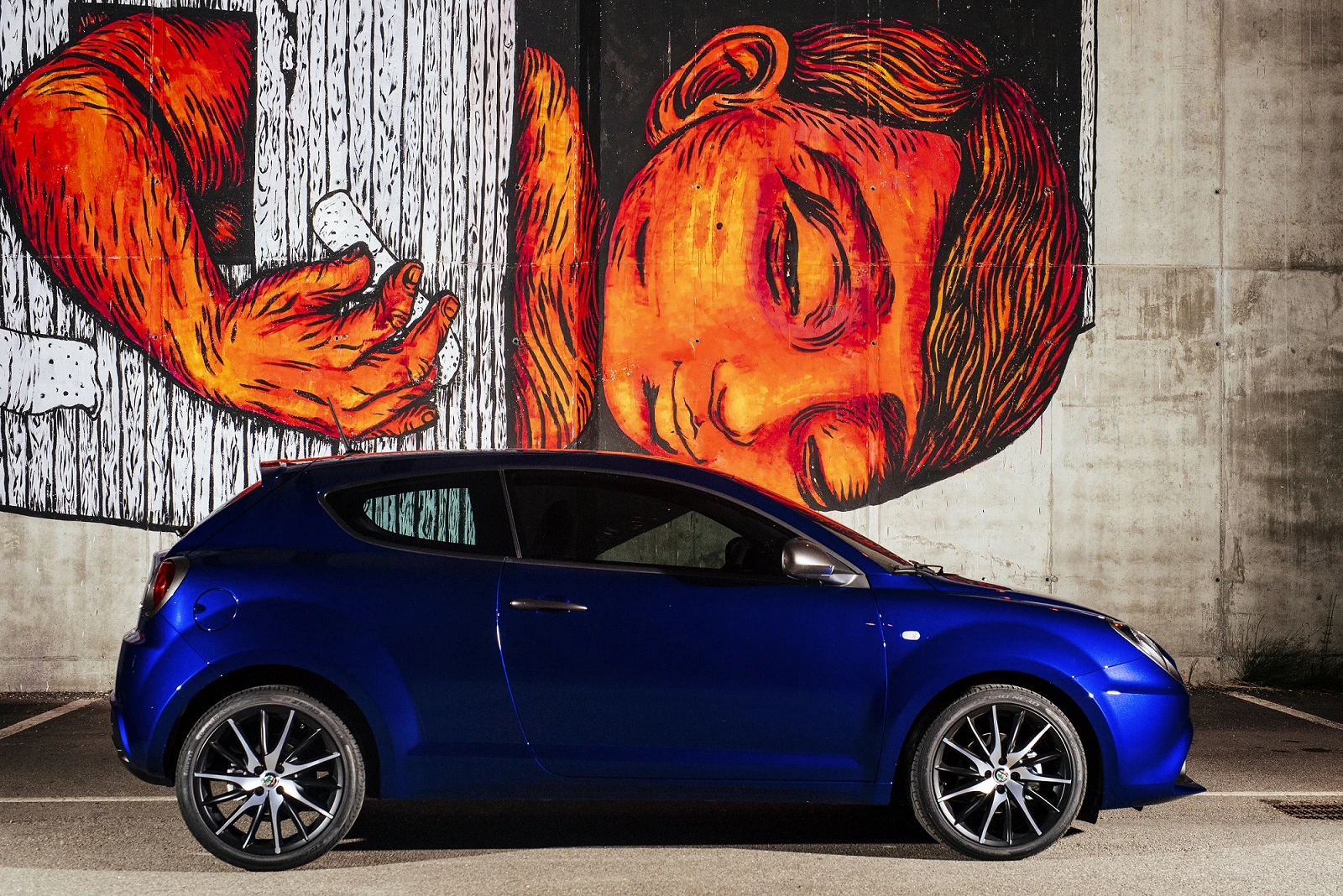Blaues Auto vor einer bemalten Wand mit einem großen, roten Gesicht eines Kindes, das die Augen geschlossen hat. Grauer Hintergrund.