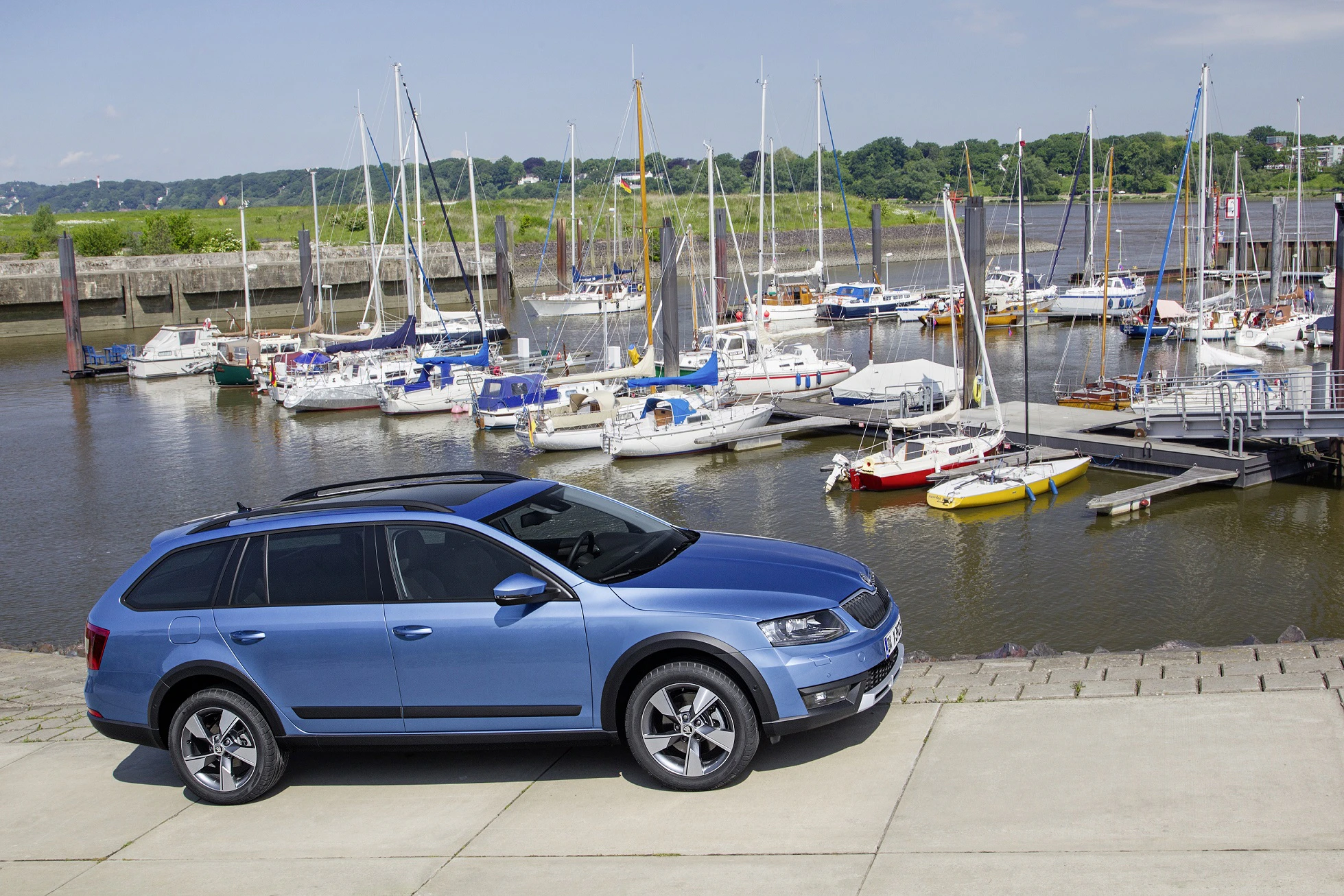 Blaues Auto parkt vor einem Yachthafen mit mehreren Segelbooten und klarem Himmel im Hintergrund.