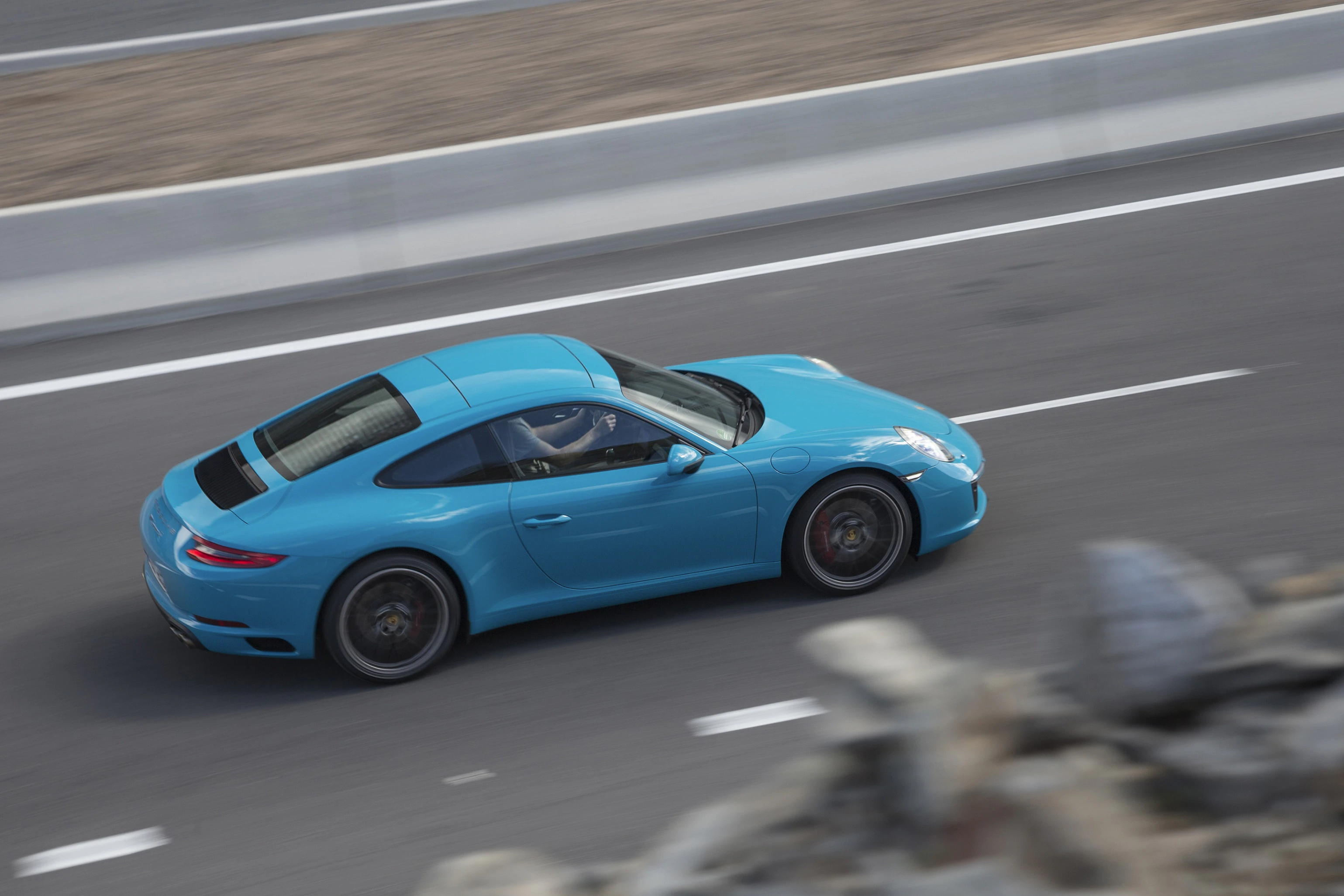 Blauer Sportwagen fährt schnell auf einer asphaltierten Straße. Aerodynamische Karosserie, modernes Design, Naturstein im Vordergrund verschwommen.