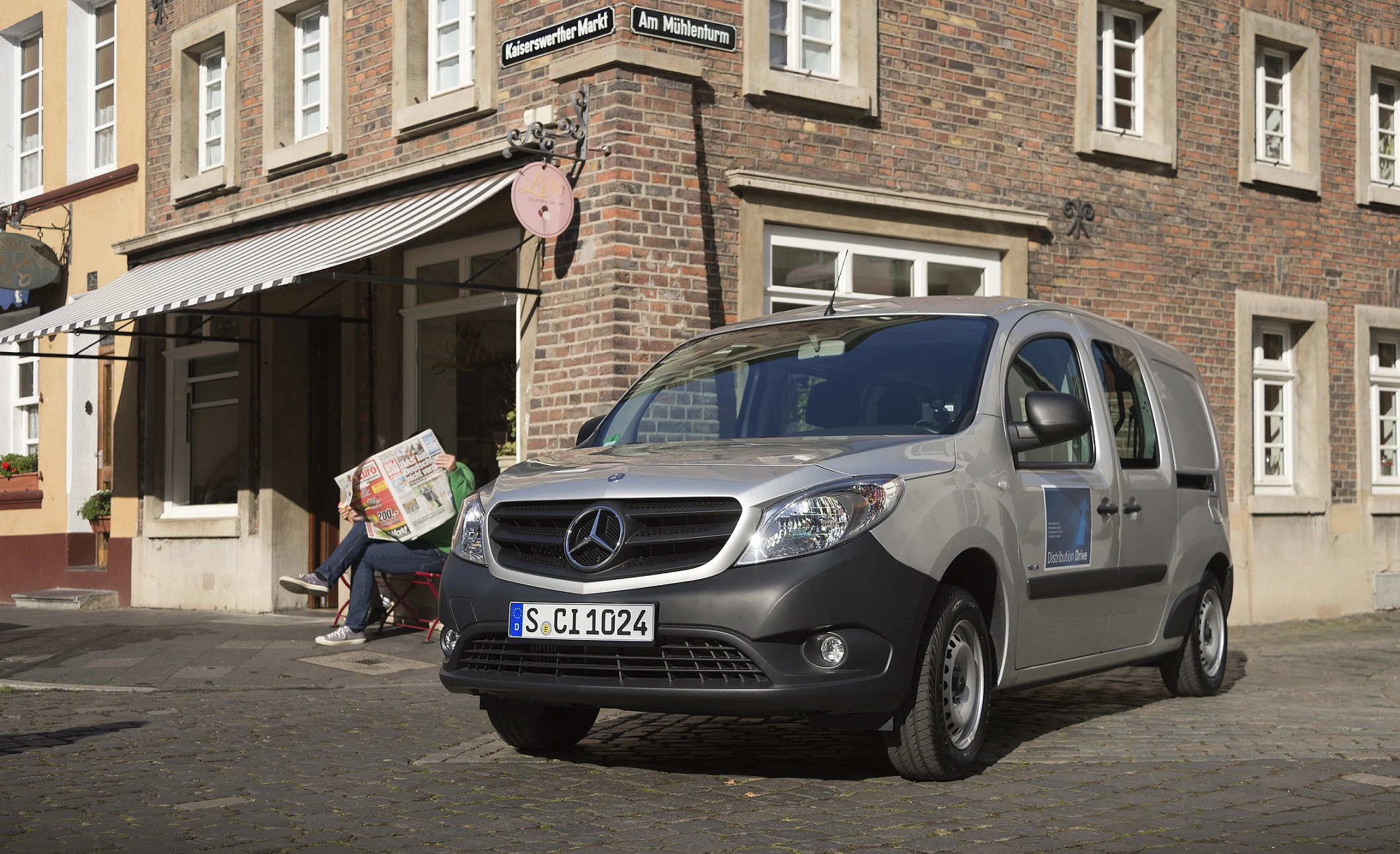 Silberner Mercedes-Van parkt auf Kopfsteinpflaster vor einem Café. Person liest Zeitung auf einer Bank vor dem Gebäude.
