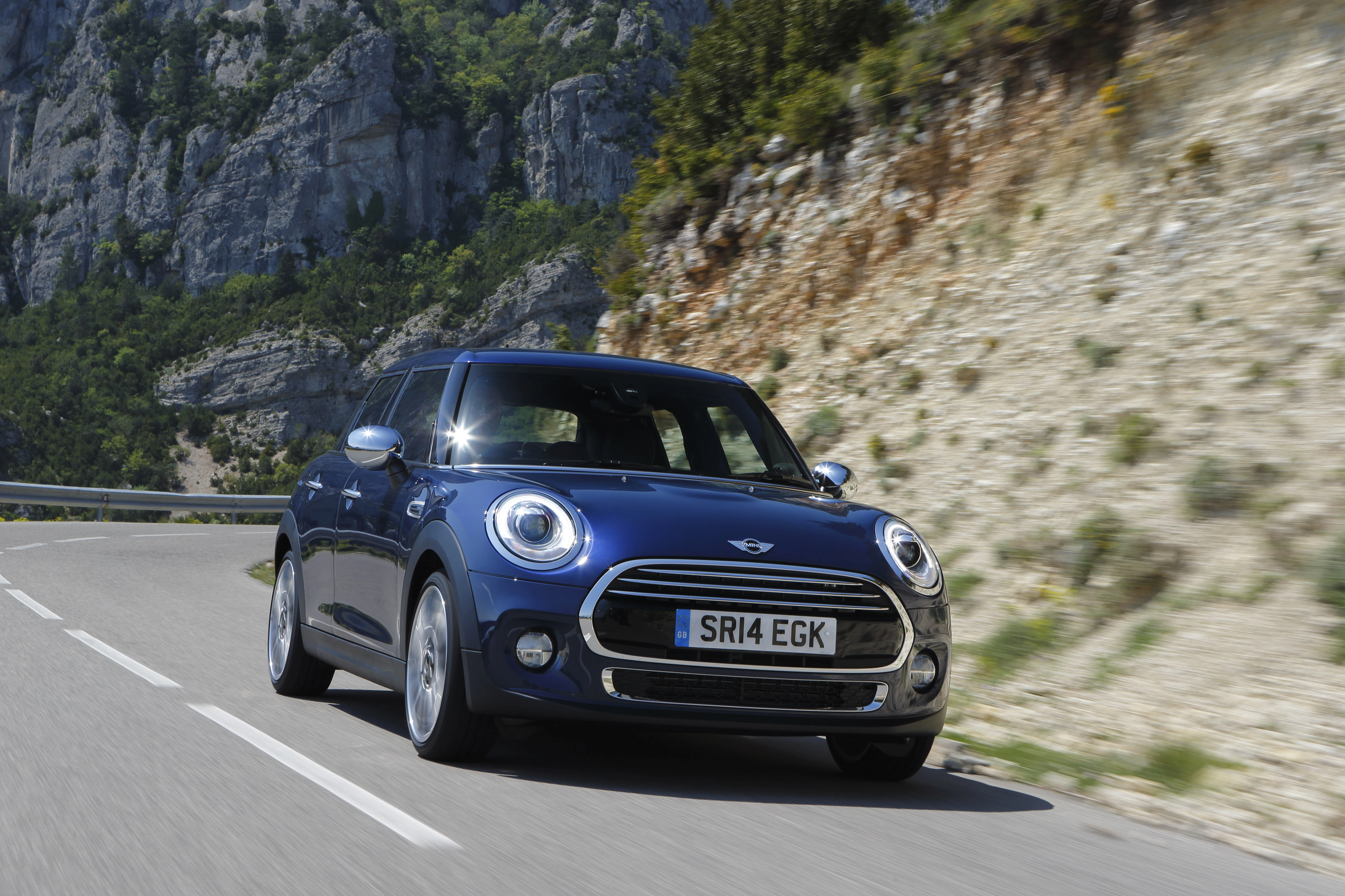 Blauer Mini Cooper fährt auf kurviger Bergstraße bei sonnigem Wetter, umgeben von Felsen und Bäumen.