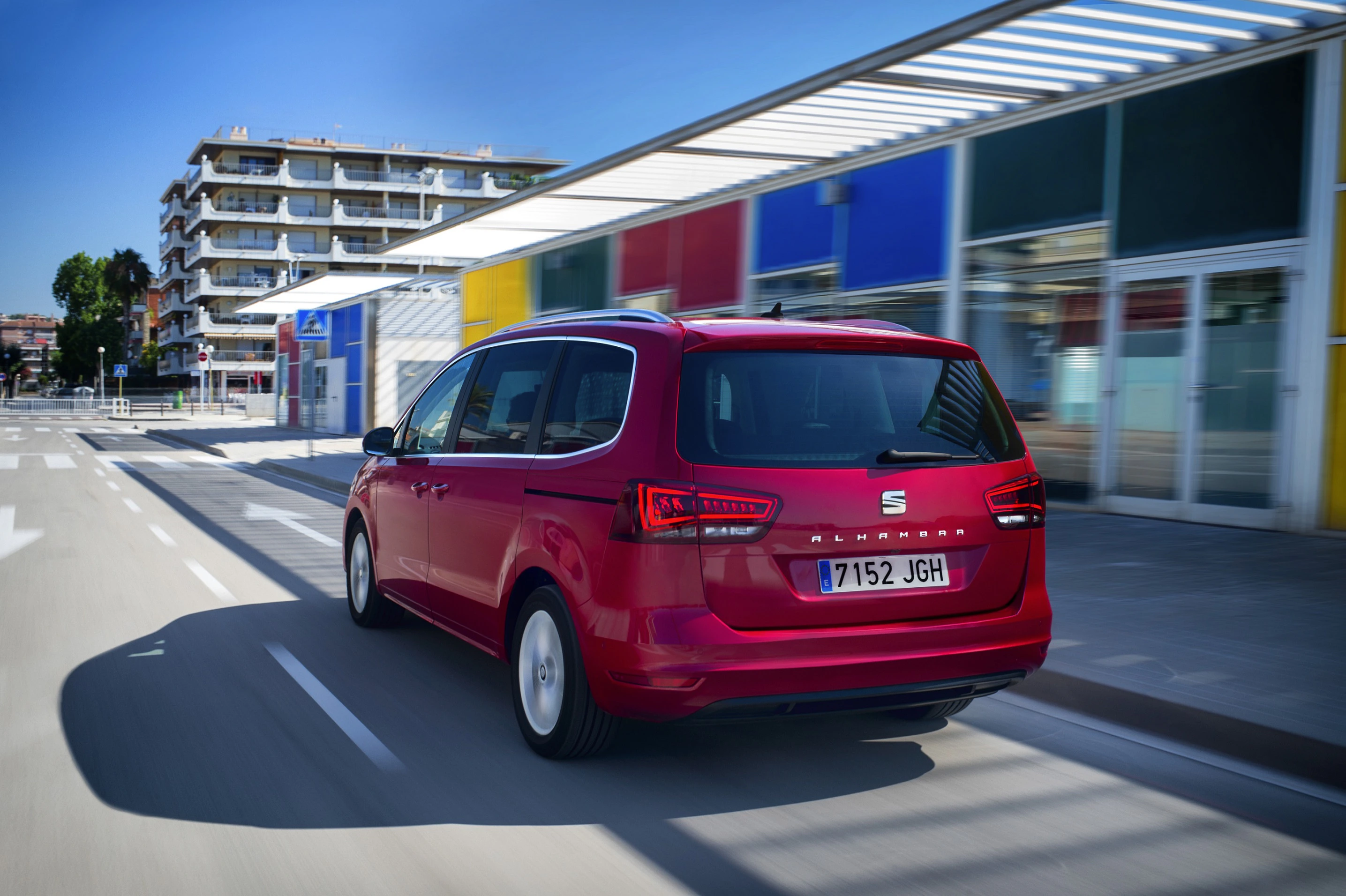 Roter Minivan fährt auf städtischer Straße, modernes Gebäude im Hintergrund, blauer Himmel, Sonnenlicht reflektiert auf dem Auto.