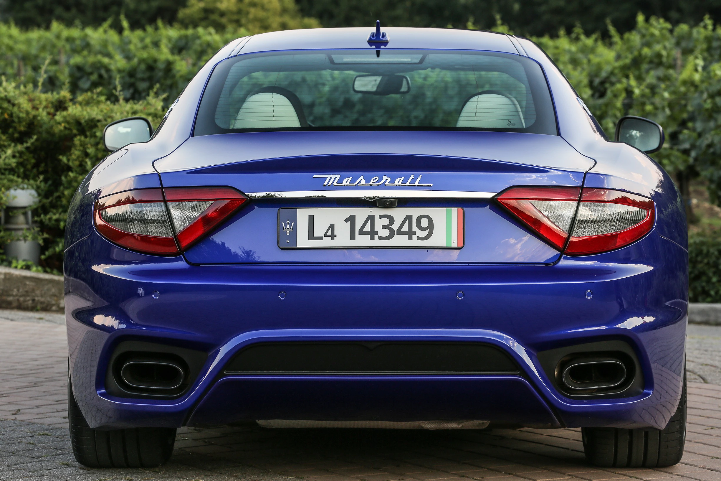 Blauer Maserati mit italienischem Kennzeichen, geparkt auf einer Pflasterstraße. Hintergrund aus Bäumen und dichter Vegetation.