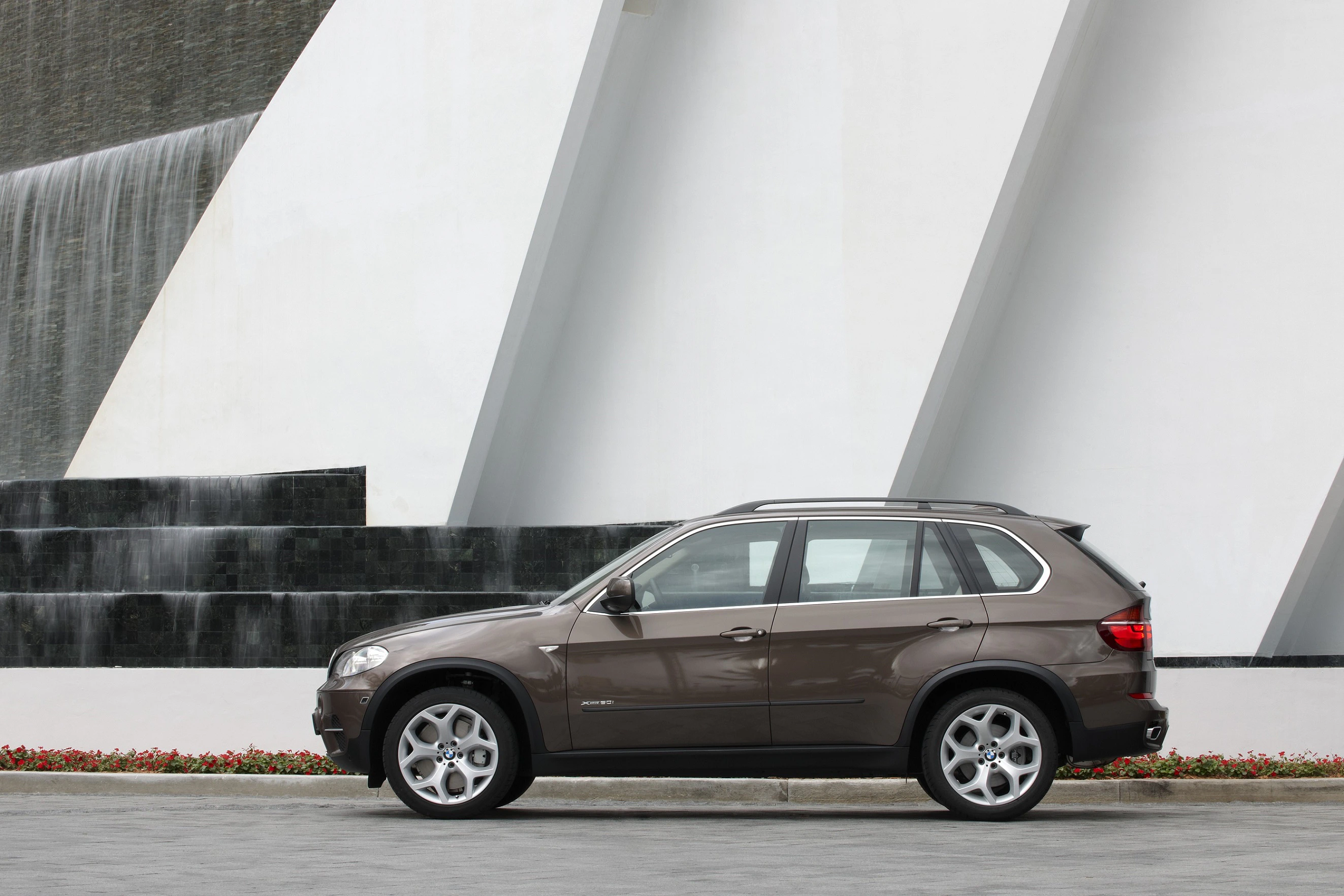 Braunes SUV parkt vor moderner Architektur mit weißer Fassade und Wasserfall; seitliche Ansicht des Fahrzeugs.