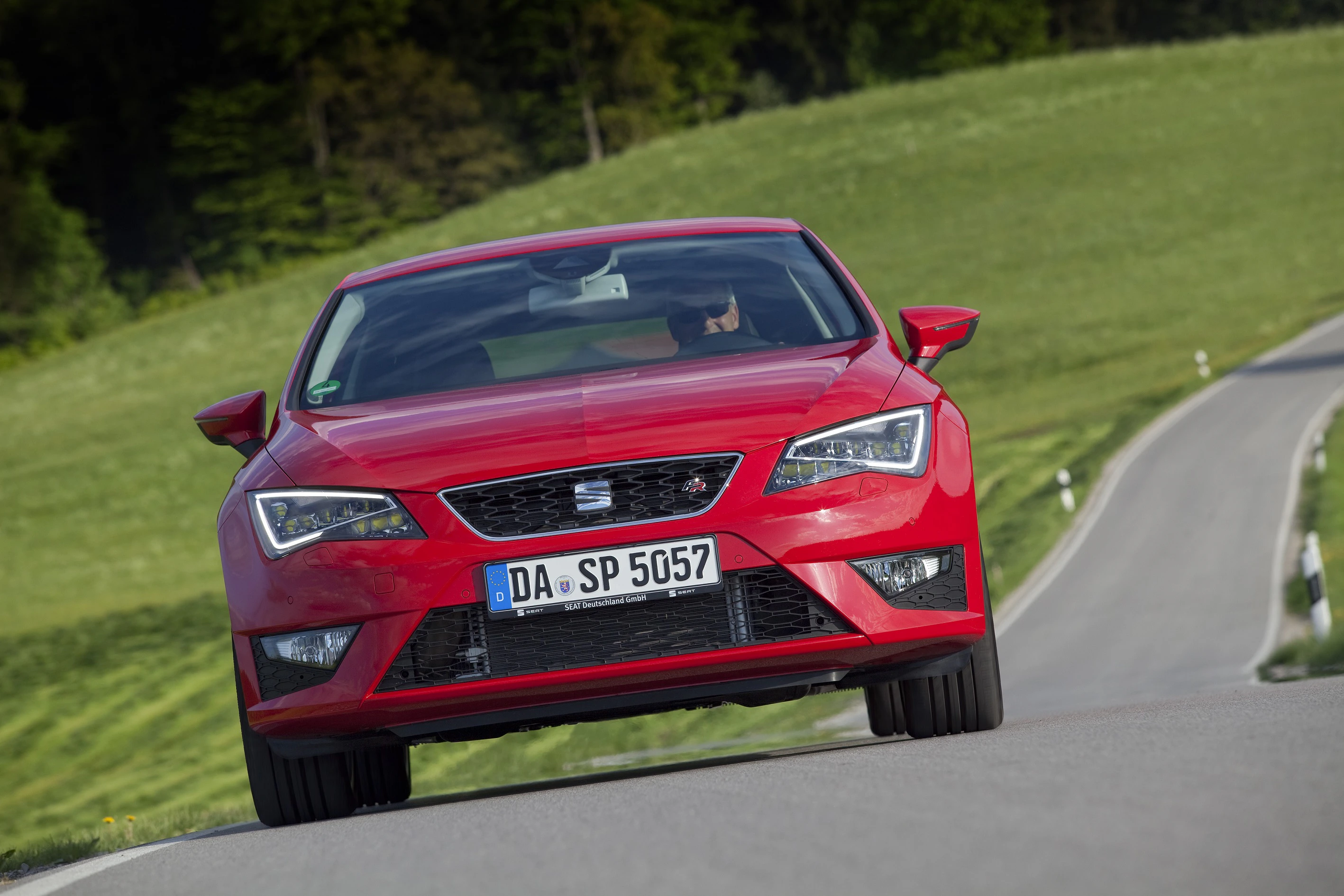 Roter Seat Leon fährt auf schmaler, gewundener Landstraße durch grüne Landschaft. Fahrer sichtbar, Auto von vorne aufgenommen.