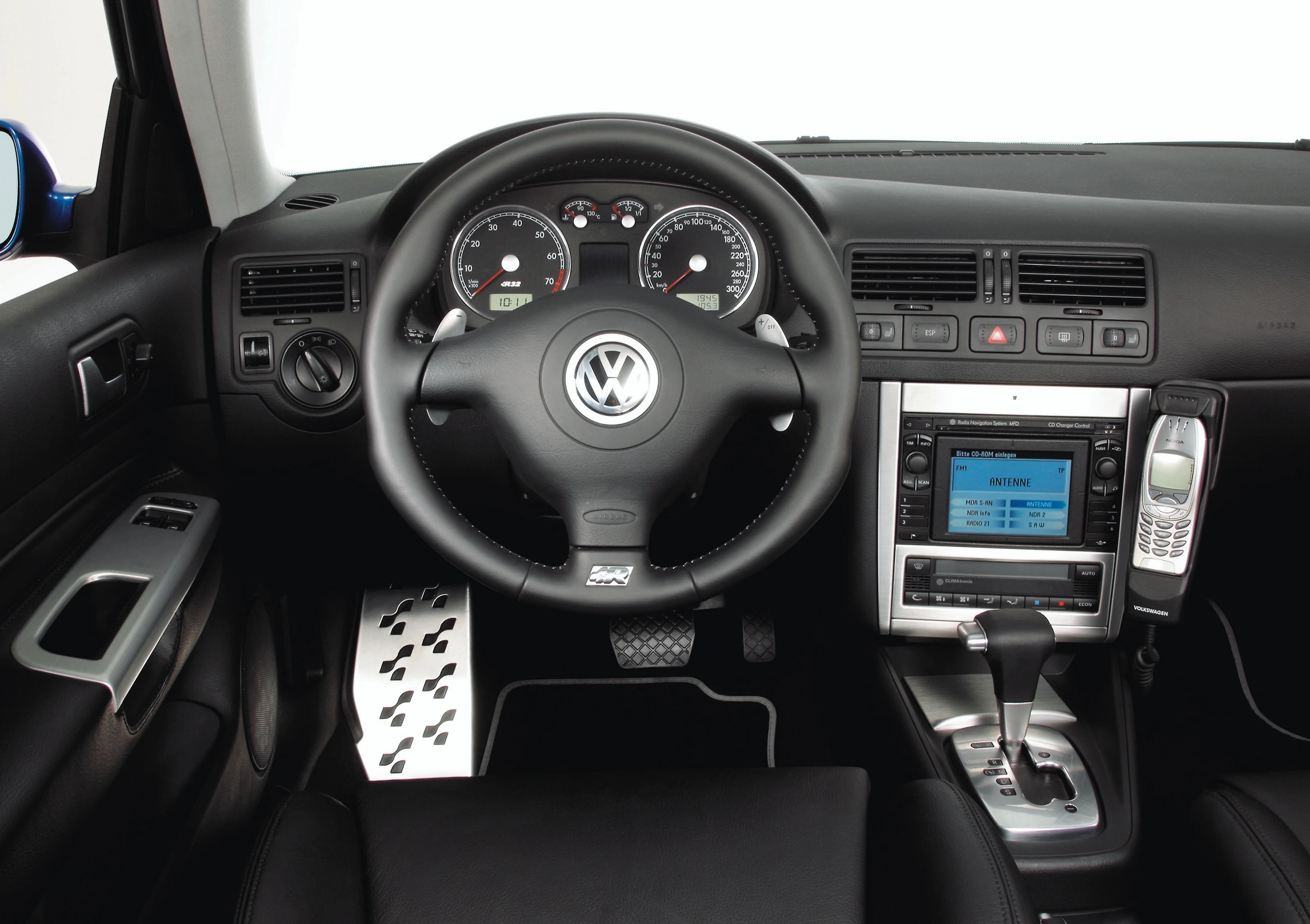 Innenraum eines VW mit Lederlenkrad, Armaturenbrett, eingebautem Bildschirm sowie Telefonhalterung und Pedale aus Metall.