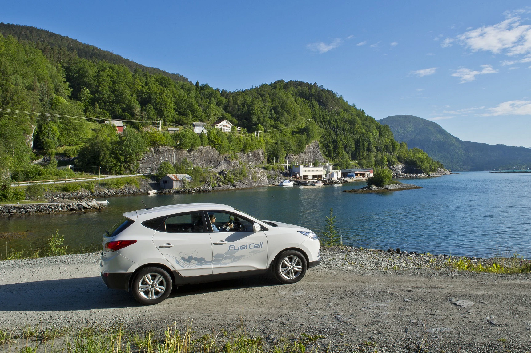Ein weißes Auto mit "Fuel Cell"-Aufschrift steht an einem Seeufer, umgeben von Bäumen und Hügeln unter blauem Himmel.