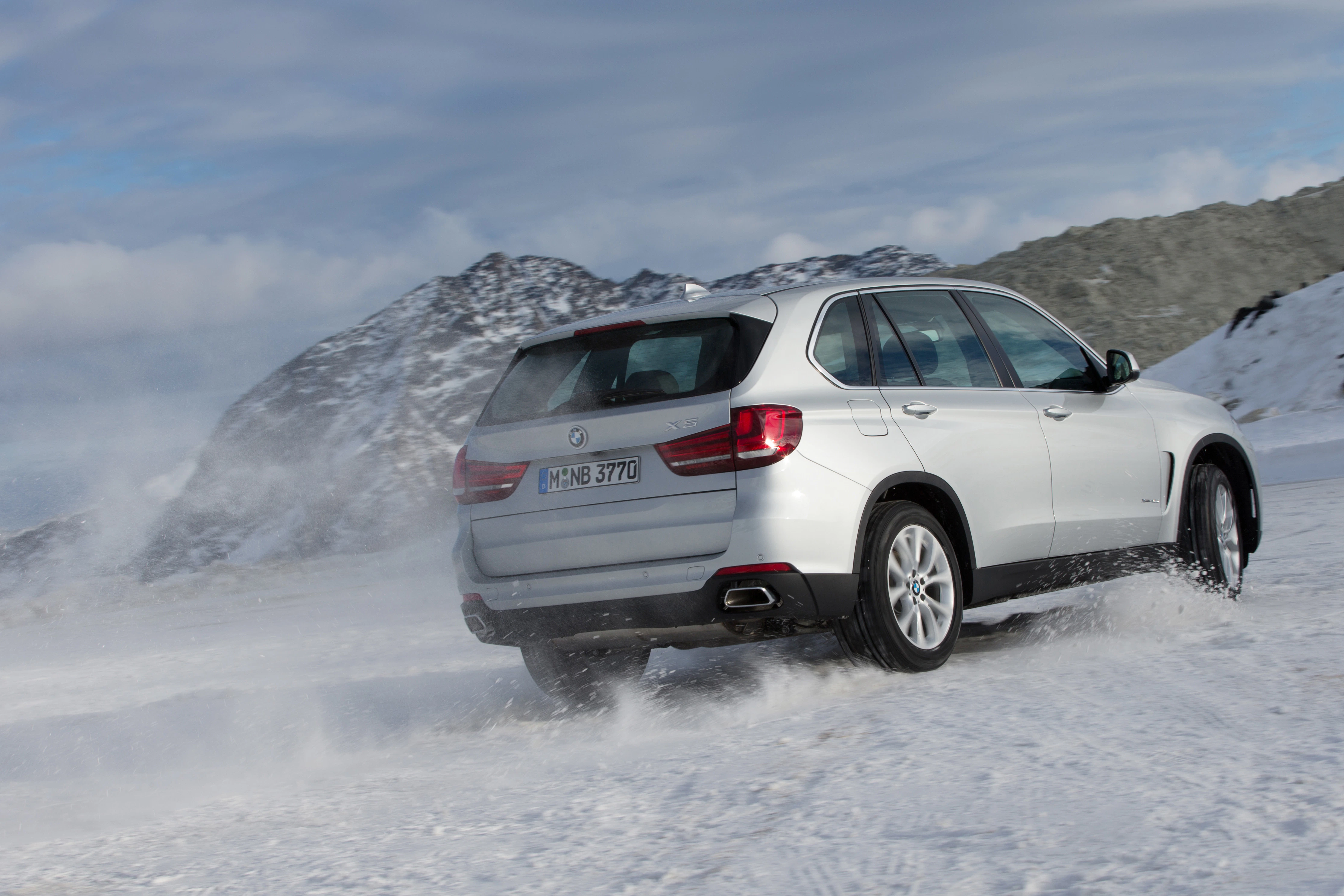 Silberner SUV fährt dynamisch auf schneebedeckter Straße in bergiger, winterlicher Landschaft. Himmel bewölkt.