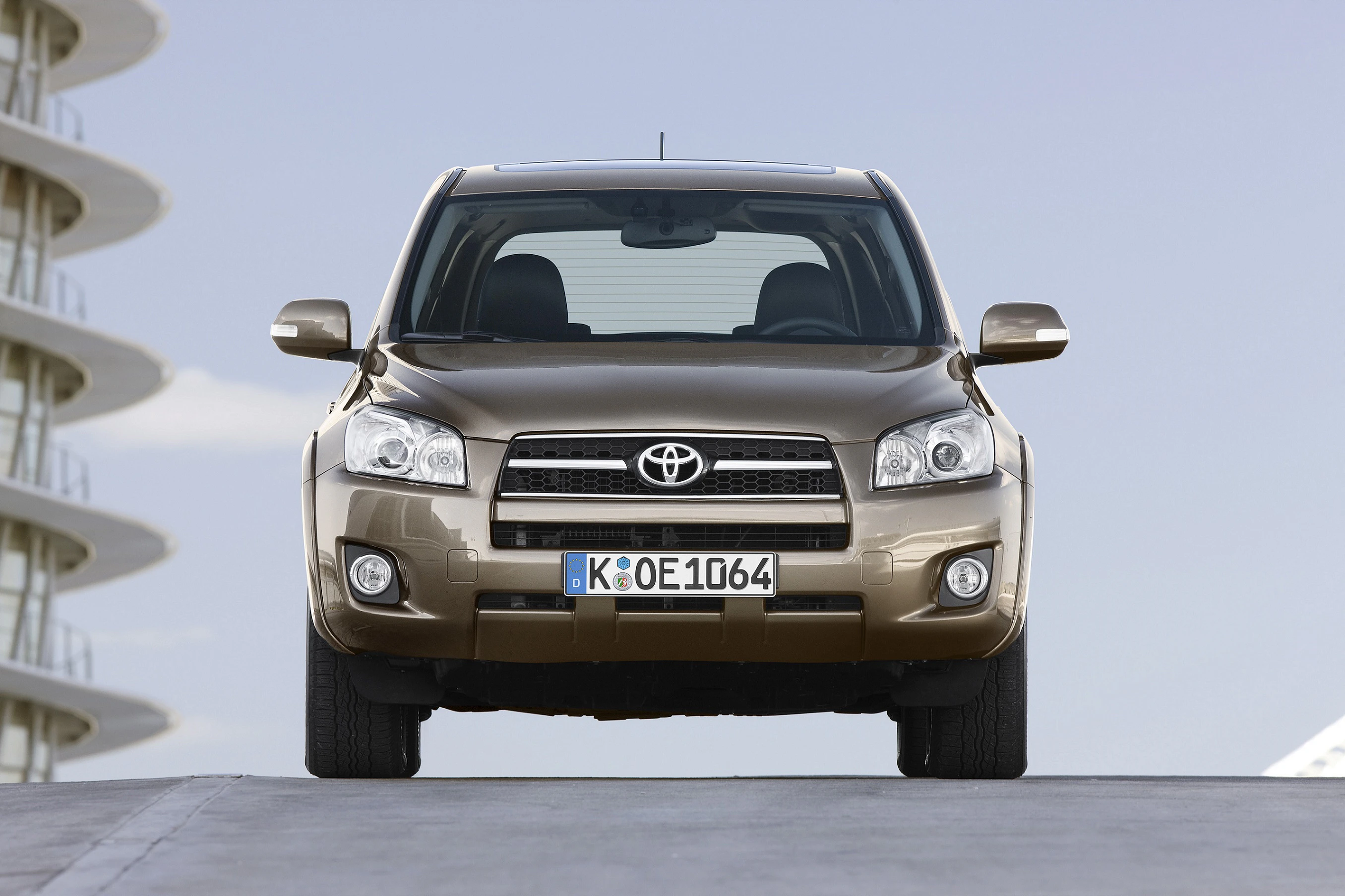 Frontansicht eines braunen Toyota SUV auf einem Betonweg vor einem modernen Gebäude mit blauen Himmel im Hintergrund.