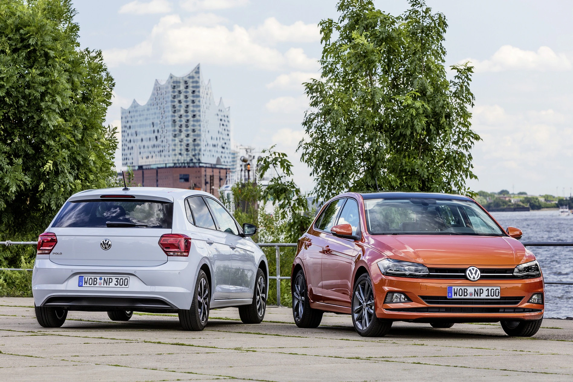 Zwei VW-Autos, ein weißer und ein orangefarbener, parken vor der Hamburger Elbphilharmonie an einem bewölkten Tag.