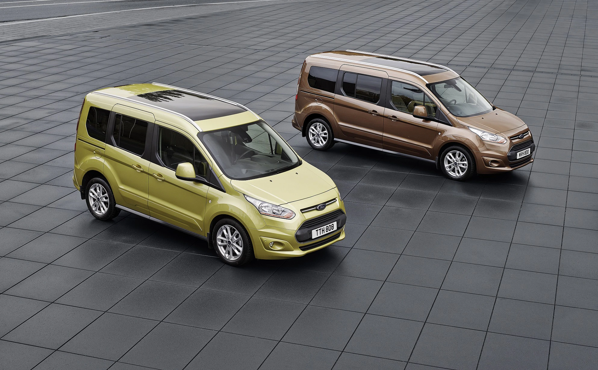 Zwei Ford Connect Vans, einer in Gelb, der andere in Braun, stehen auf einem gepflasterten Platz nebeneinander.