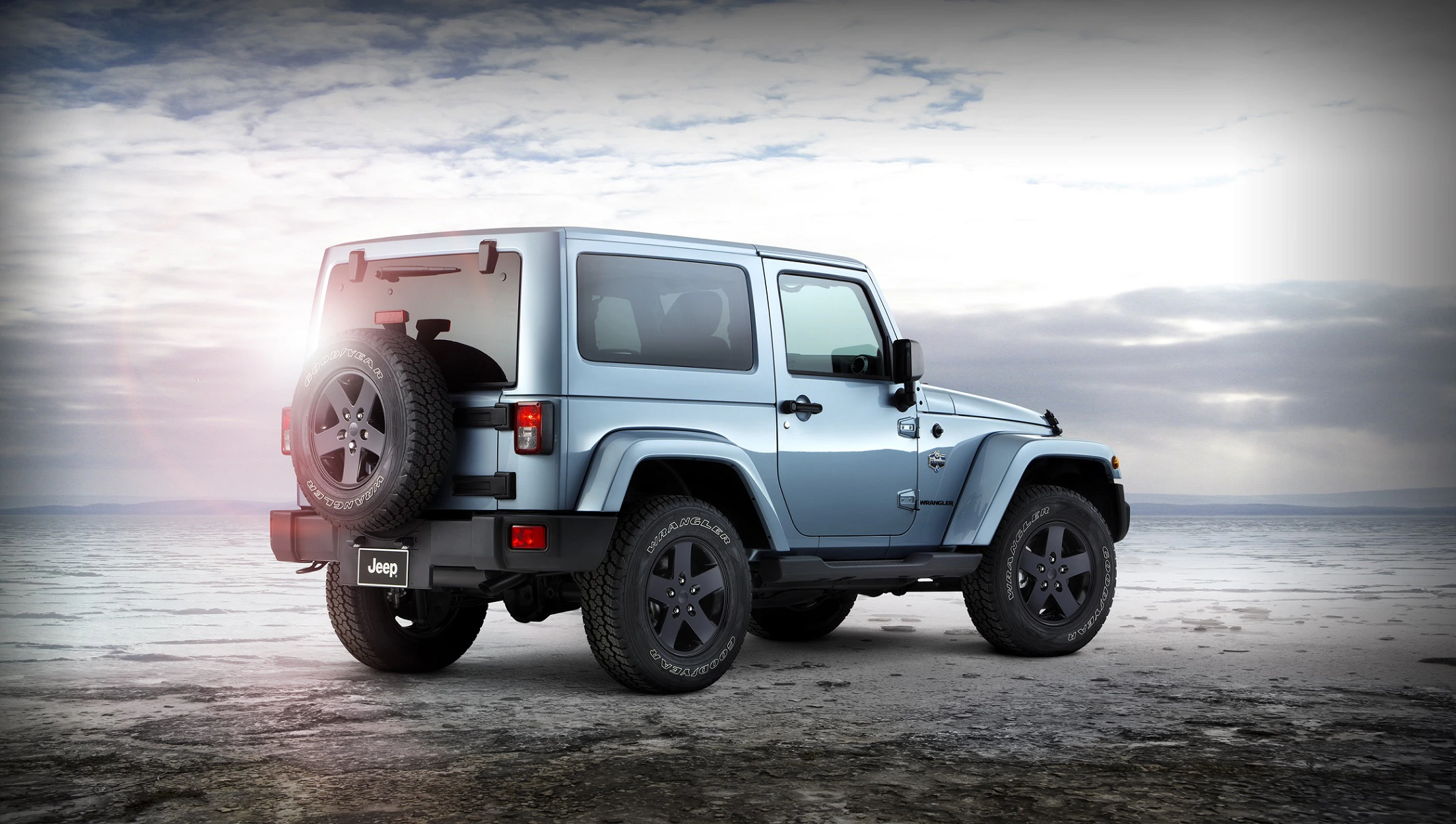Hellblauer Jeep Wrangler mit Ersatzreifen, auf trockener Ebene bei bewölktem Himmel geparkt. Rückenansicht zeigt Fahrzeugmarkenzeichen.