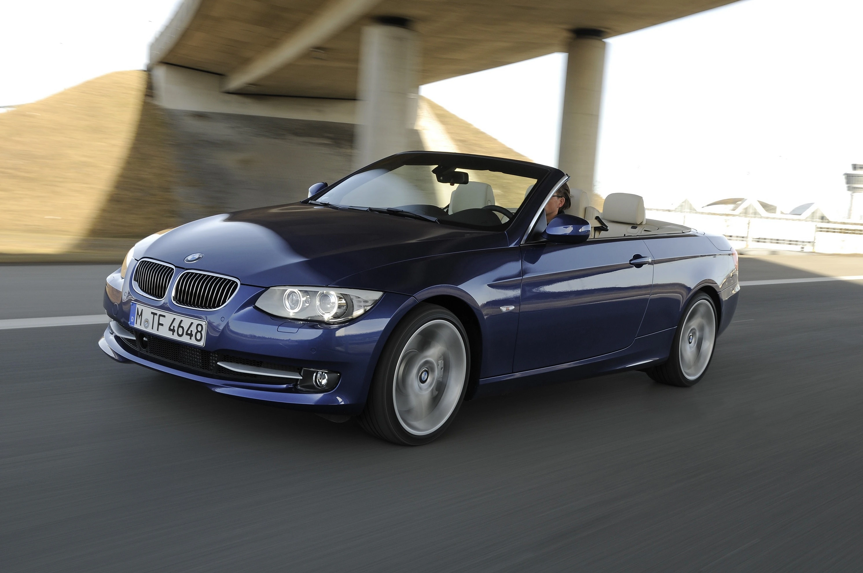 Blaues BMW Cabrio fährt unter einer Brücke auf einer Straße bei Tageslicht. Das Dach ist geöffnet, Fahrer sichtbar.