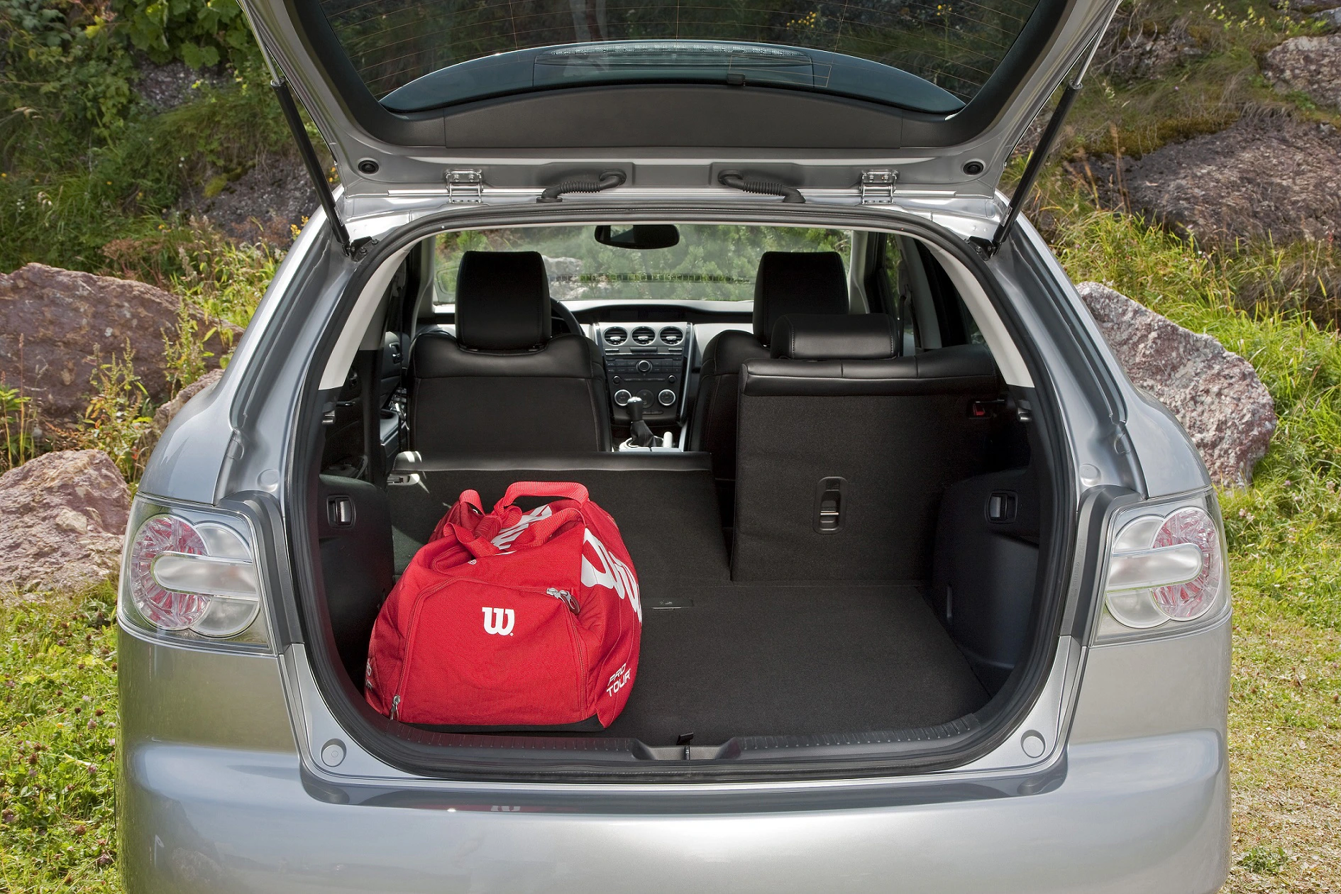 Offener Kofferraum eines silbernen Autos, mit roter Sporttasche darin. Im Hintergrund ist eine grüne Landschaft zu sehen.