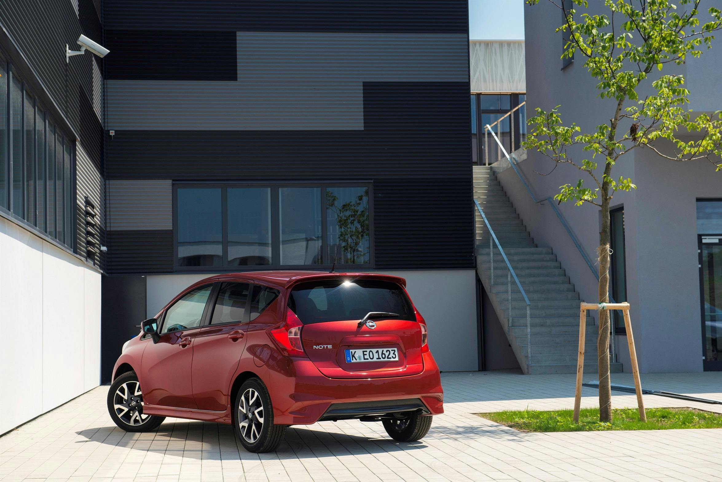 Rotes Auto parkt vor einem modernen Gebäude mit schwarzer Fassade und Treppenaufgang, ein junger Baum steht auf dem hellen Pflaster.