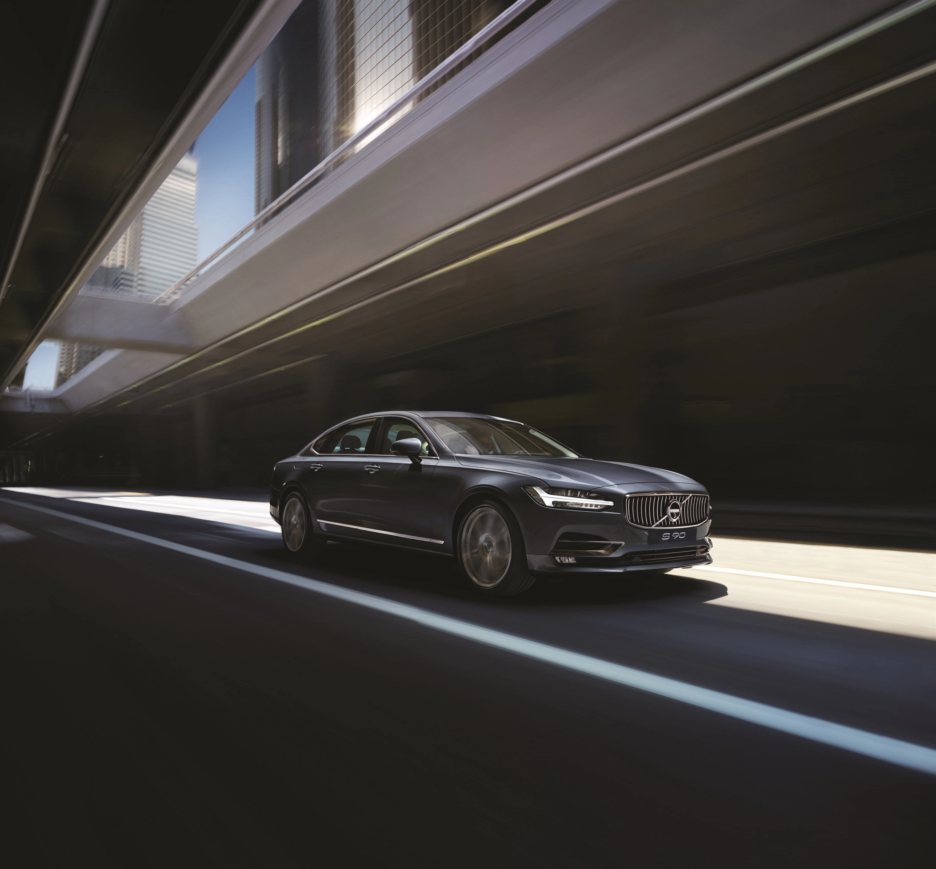 Schwarzer Volvo S90 fährt dynamisch durch einen modernen Tunnel unter Hochhauskomplexen bei Tageslicht.